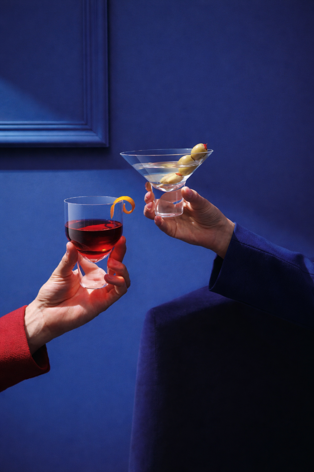 Two people are holding drinks; one has a glass of red wine with a twist of orange peel, and the other has a martini glass with green olives. The background is blue.