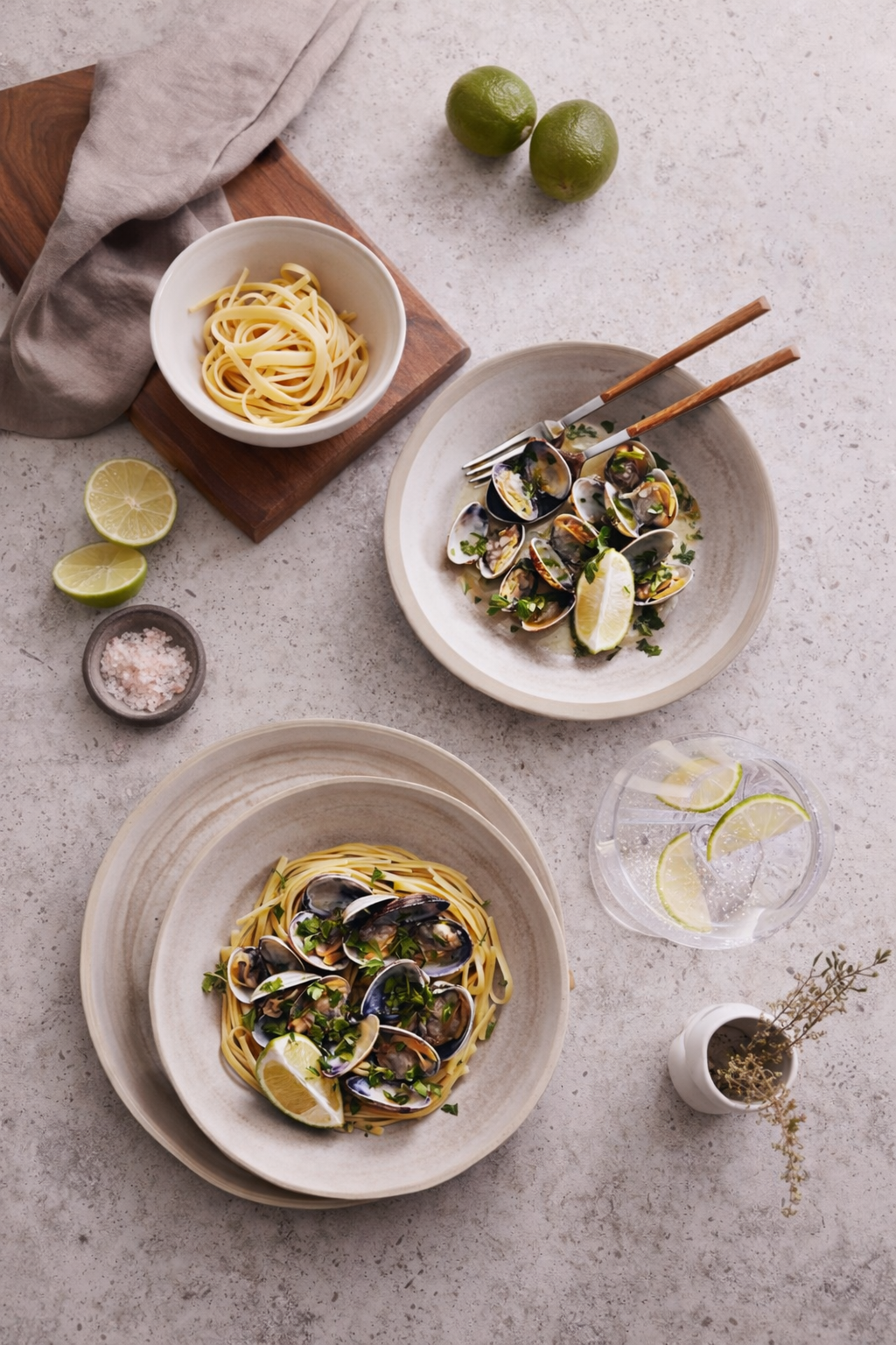 A flat lay of two plates of linguine with mussels garnished with herbs and lime wedges, a bowl of cooked linguine, a bowl of raw mussels, lime halves, a small dish of sea salt, a glass of water with lime slices, two green limes, and small sprigs of h