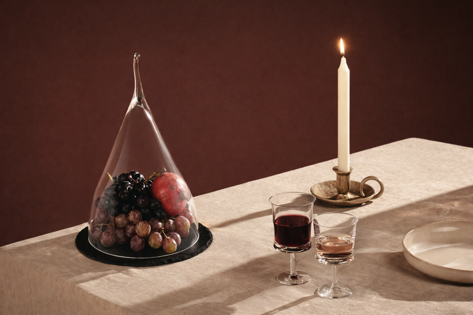 A table with a glass cloche covering grapes, an apple, and a small bunch of grapes; two glasses of red wine; an empty glass; a lit candle in a brass holder; a white ceramic plate; all on a beige tablecloth against a dark background.