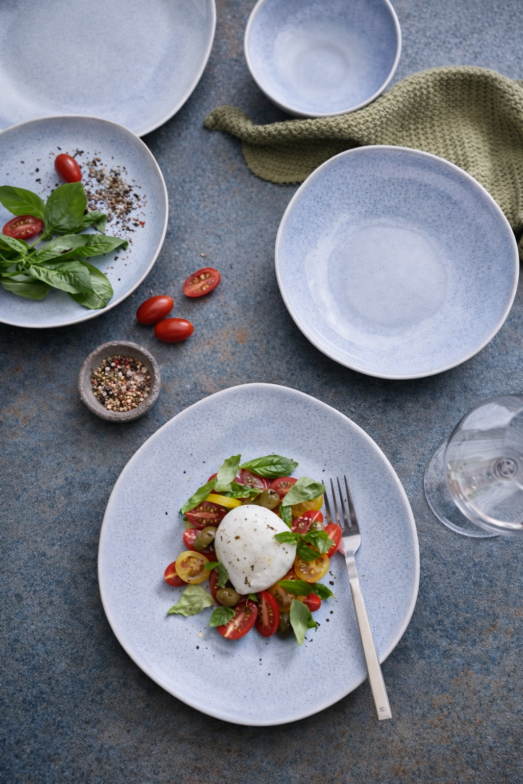 A partially eaten Caprese salad on a white speckled plate with cherry tomatoes, fresh basil, and mozzarella, set on a blue textured table with empty blue bowls, a cloth napkin, a glass of water, and some scattered cherry tomatoes.