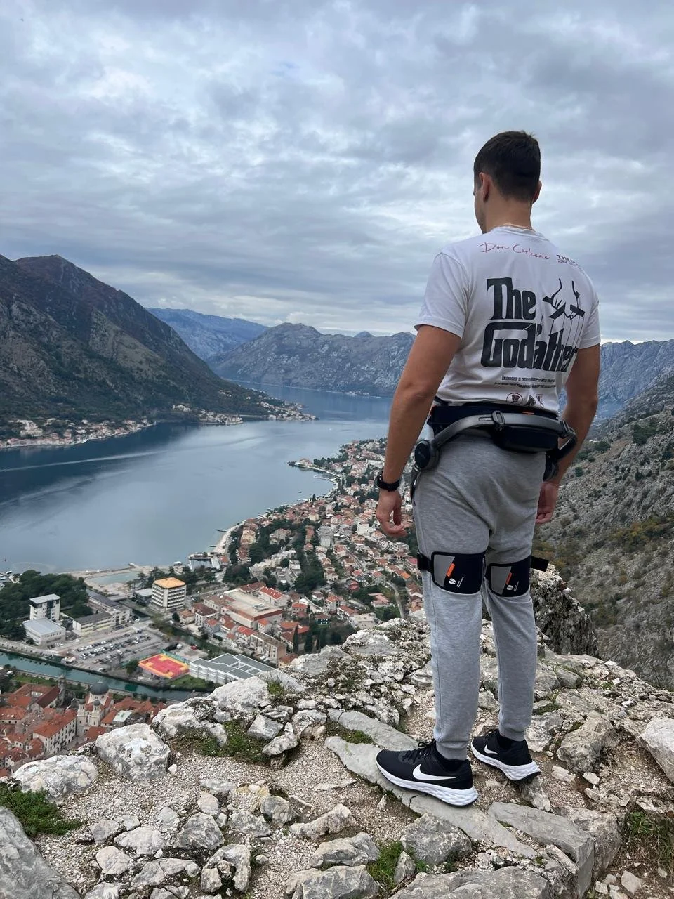 Man standing on rocky ledge overlooking a valley with a river, surrounded by mountains, in cloudy weather.