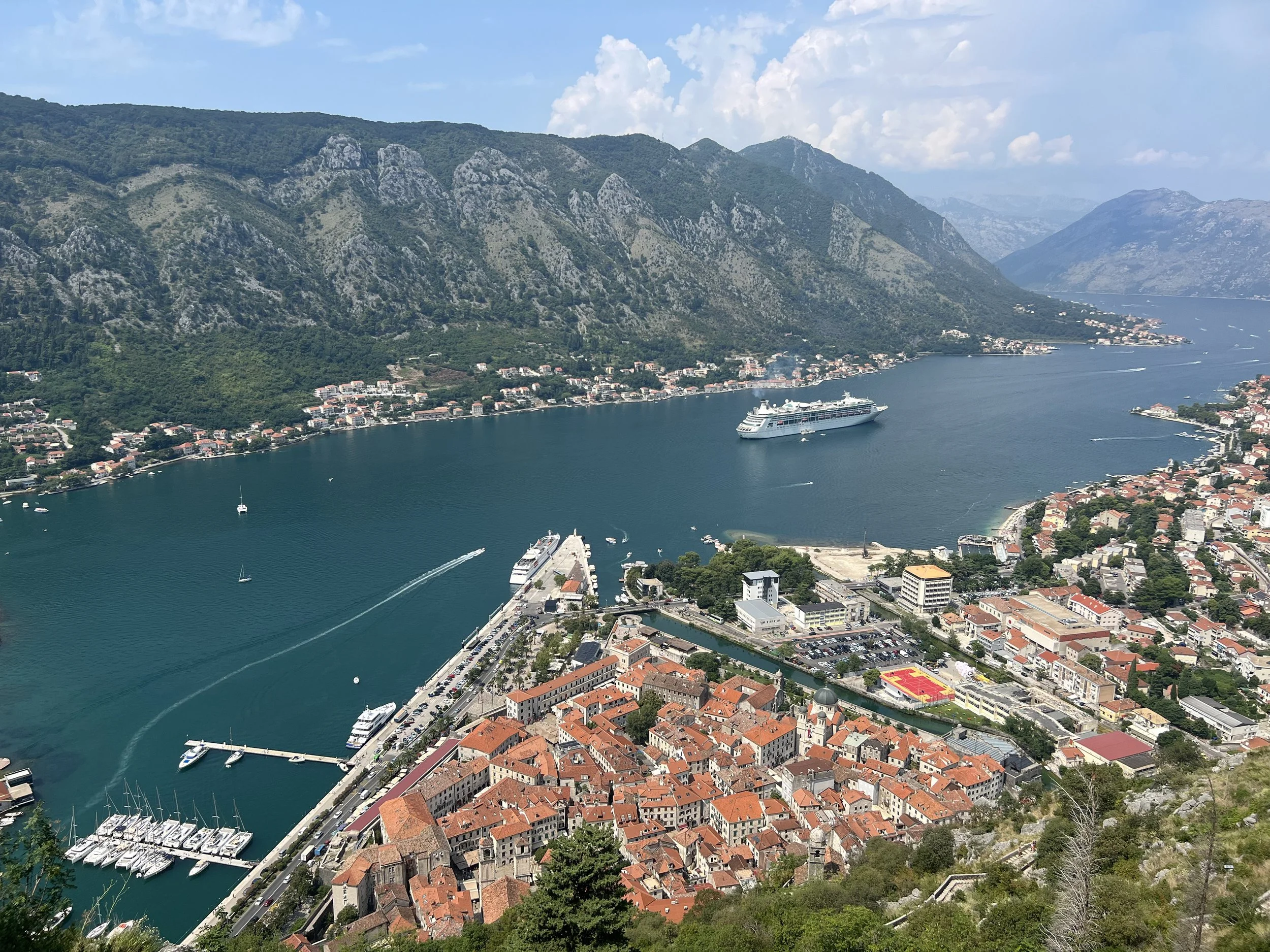 Aerial view of a coastal town with red rooftops, a marina with boats, a large cruise ship in the bay, surrounded by green mountains and a body of water.