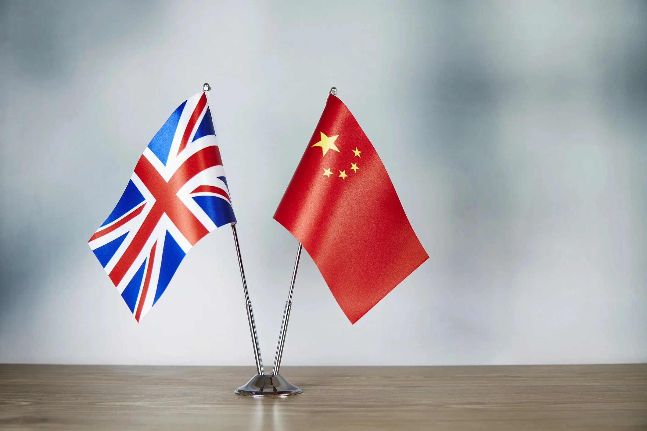 Two small flags, one of the United Kingdom and the other of China, displayed on metal sticks in a silver stand on a wooden surface against a plain light background.