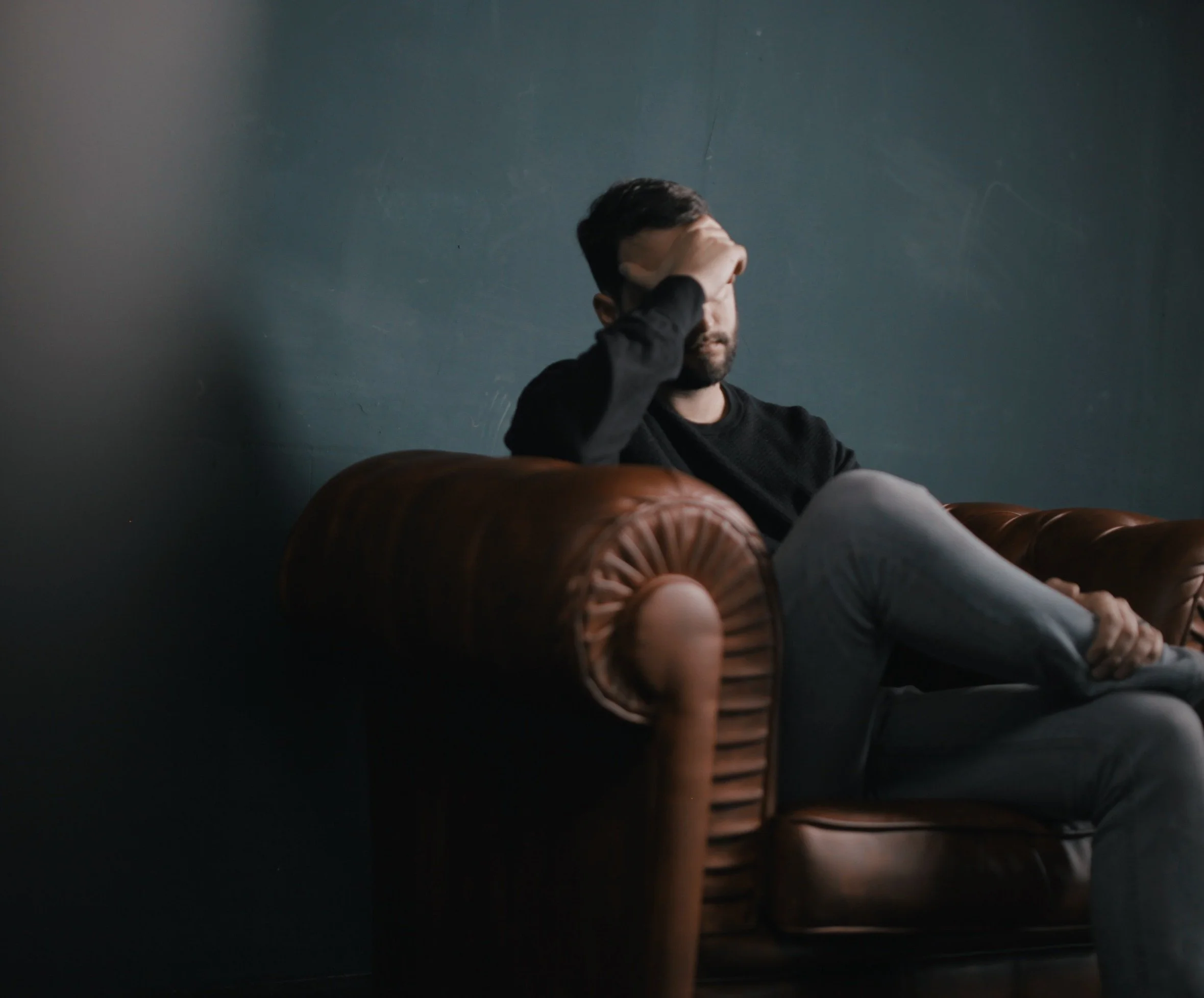 A man sitting on a leather sofa, holding his forehead with one hand, appearing stressed or deep in thought, against a dark, plain wall.