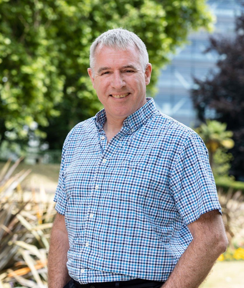 Smiling middle-aged man with short gray hair, wearing a blue and white checkered shirt, standing outdoors with greenery and trees in the background.