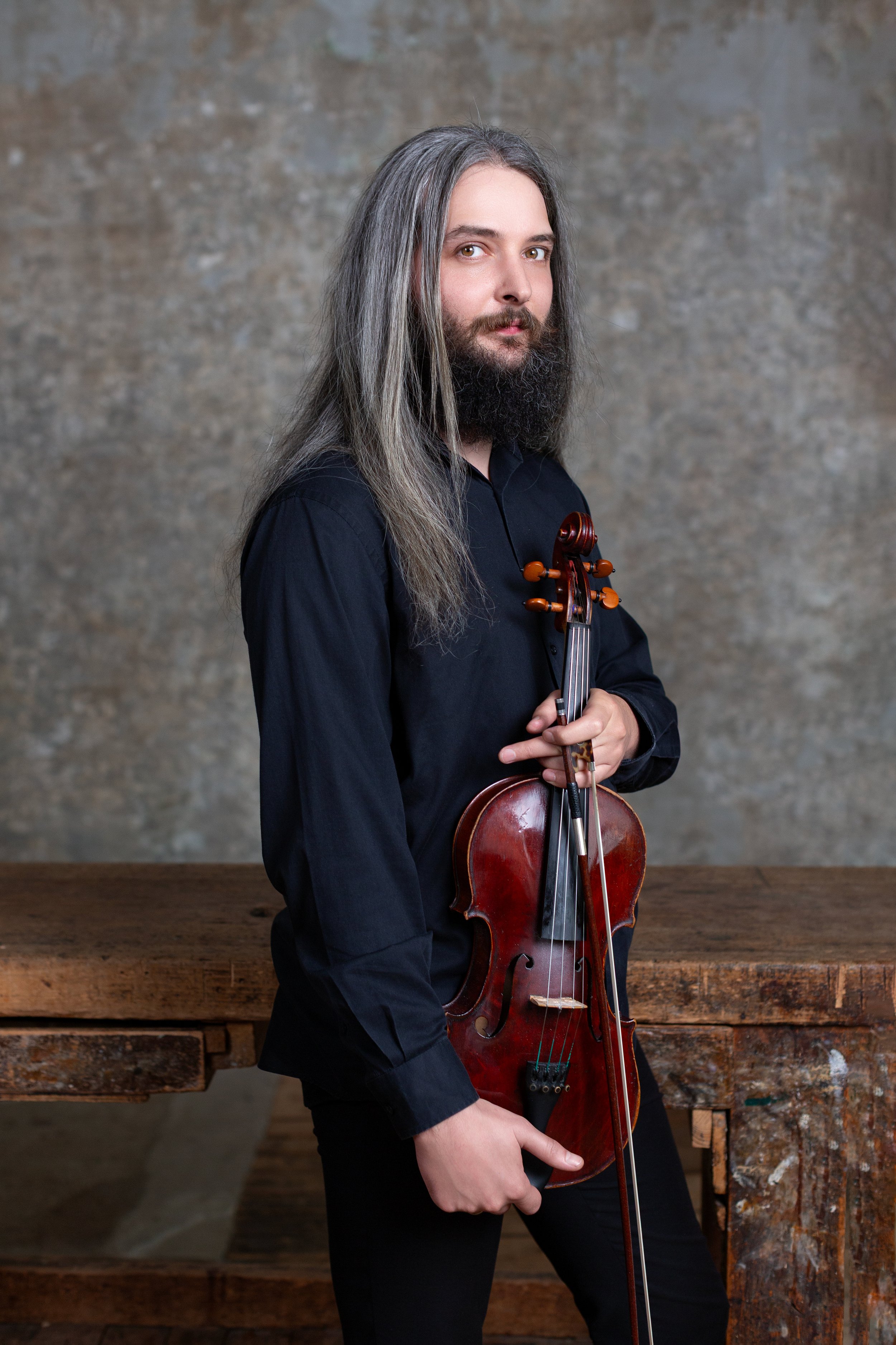Un homme avec une barbe et cheveux longs tenant un violon, portant une chemise noire, devant une surface en pierre et un meuble en bois.