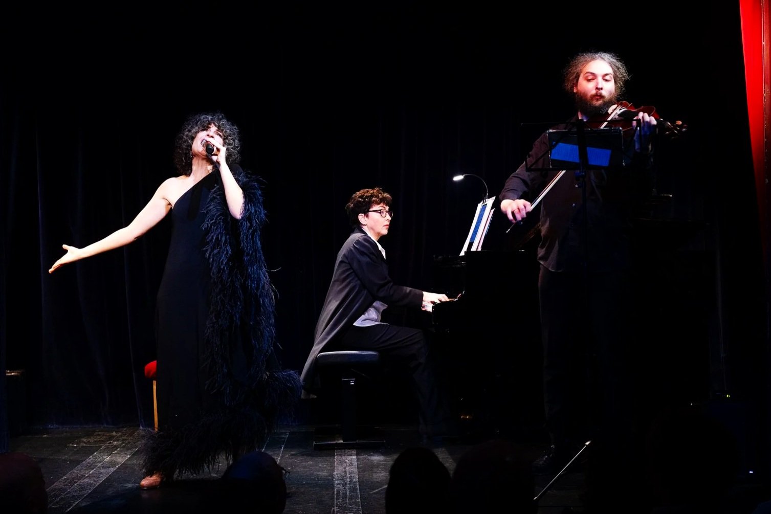 Trois musiciens en performance sur scène, une chanteuse élégante en robe noire avec une veste en plumes, une pianiste et un violoniste, en noir, dans une ambiance de concert. Theather Yiddish