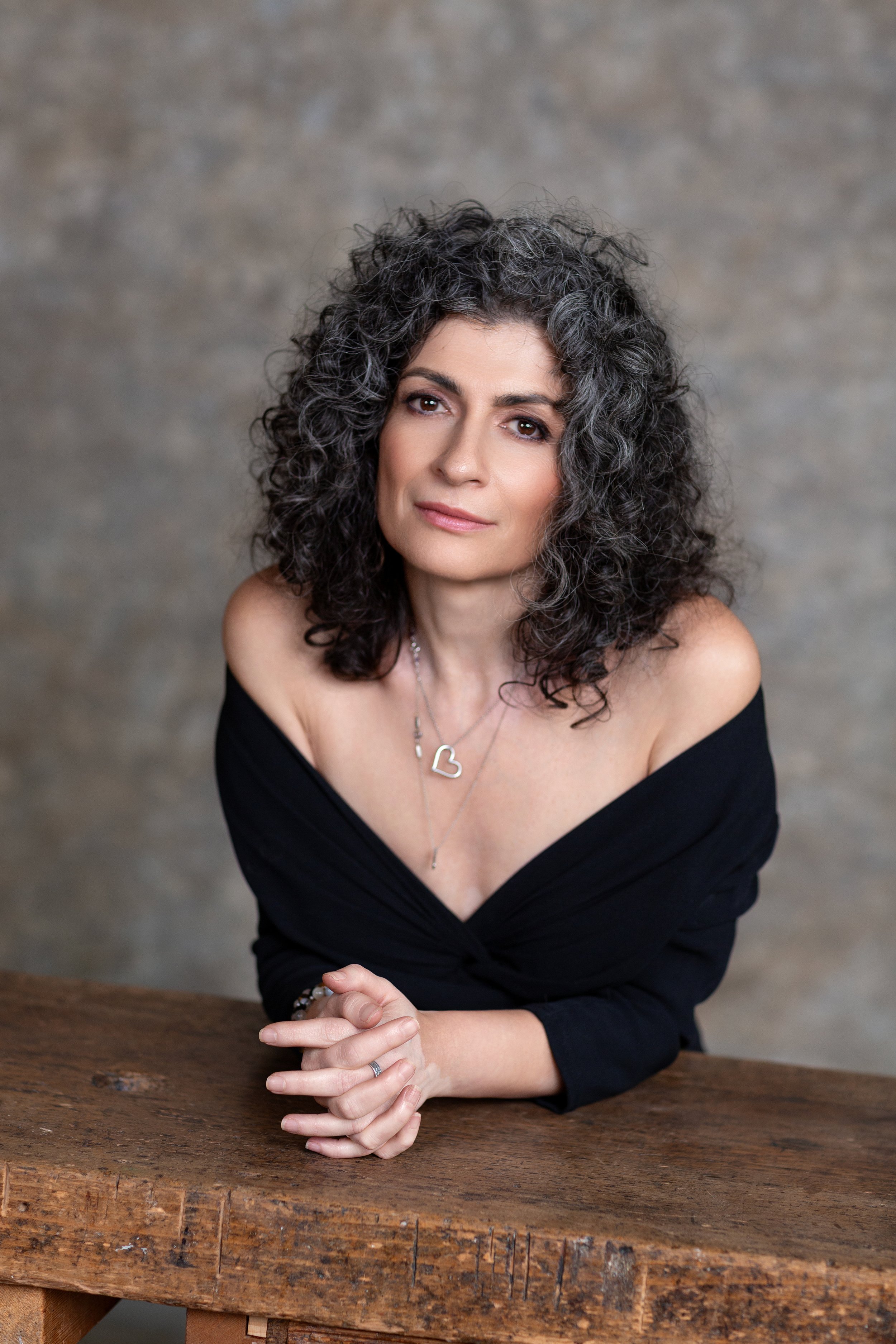 Une femme aux cheveux gris et bouclés, portant un haut noir dénudant les épaules, assise à une table en bois, regardant vers l'appareil photo, avec une touche élégante de bijoux.