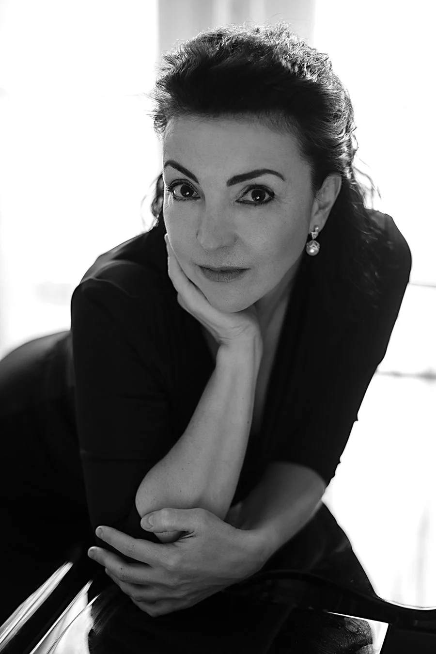 Portrait noir et blanc d'une femme avec des cheveux foncés, portant des boucles d'oreilles en perle, posant la main sur son visage, avec un sourire doux