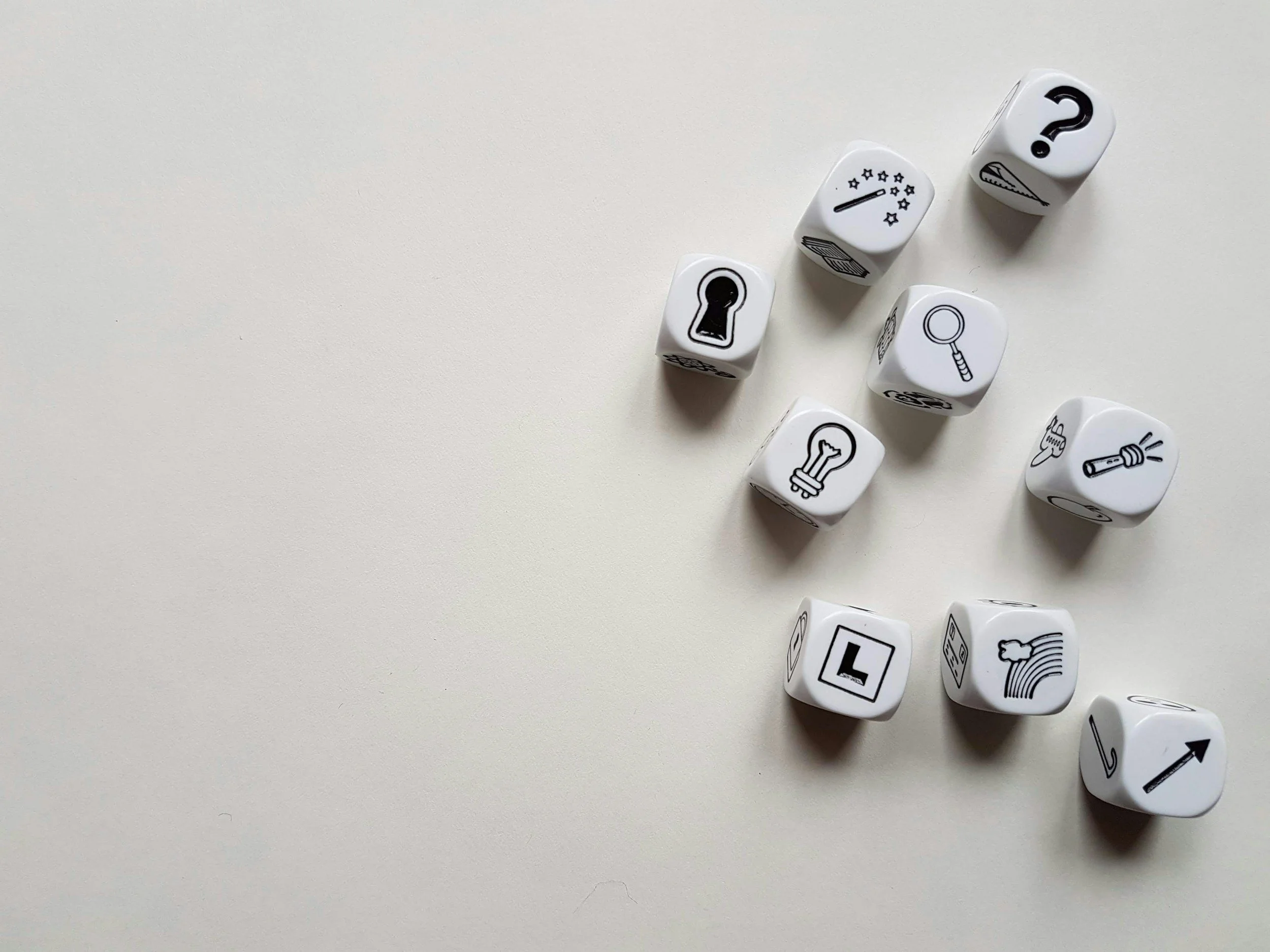 A set of nine white dice with black symbols, including a keyhole, light bulb, magnifying glass, lightning bolt, question mark, streaks, arrow, and other icons, scattered on a light-colored surface.
