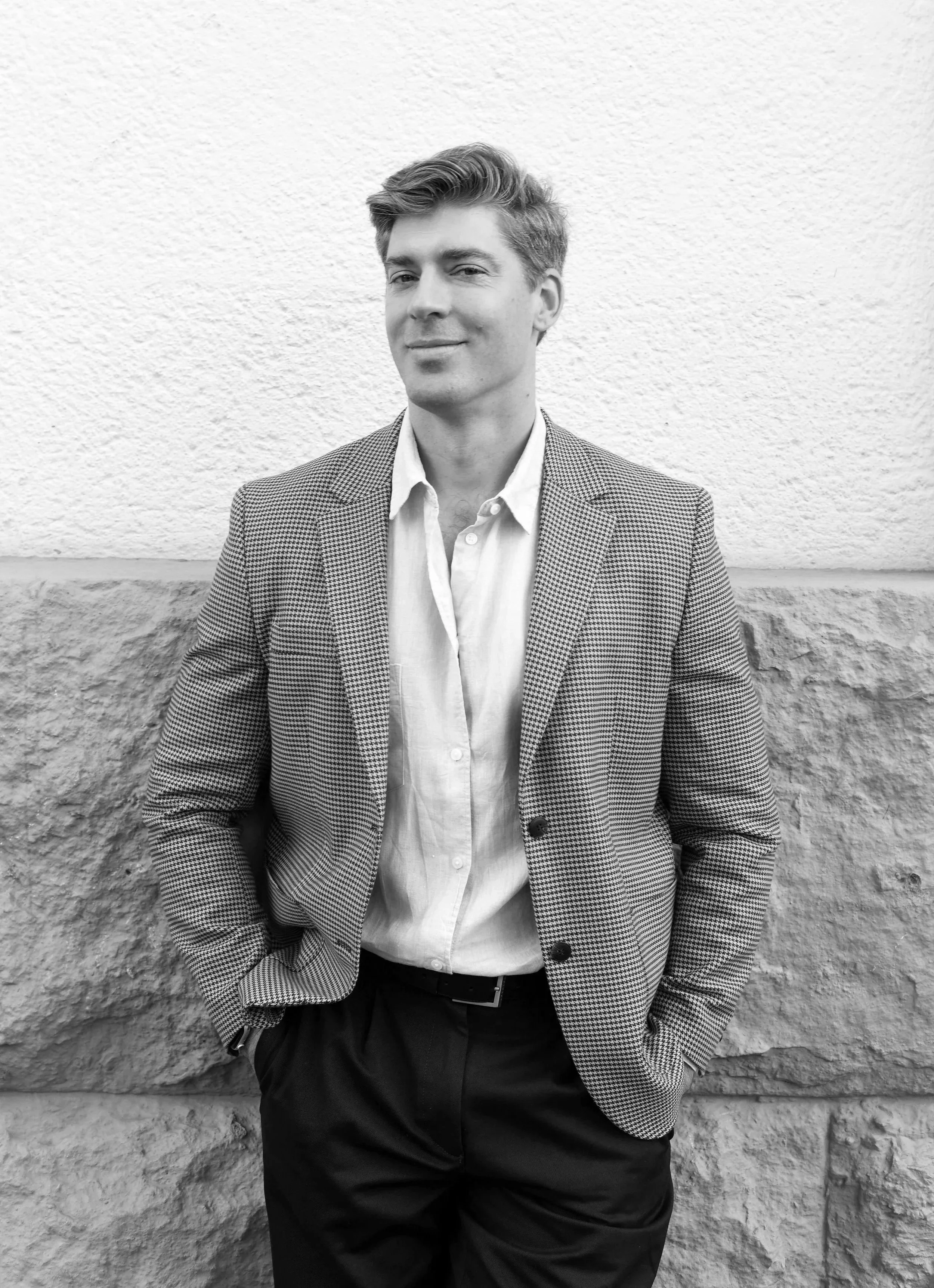 A young man in a checkered blazer and white shirt stands against a textured wall, hands in his pockets, smiling slightly.