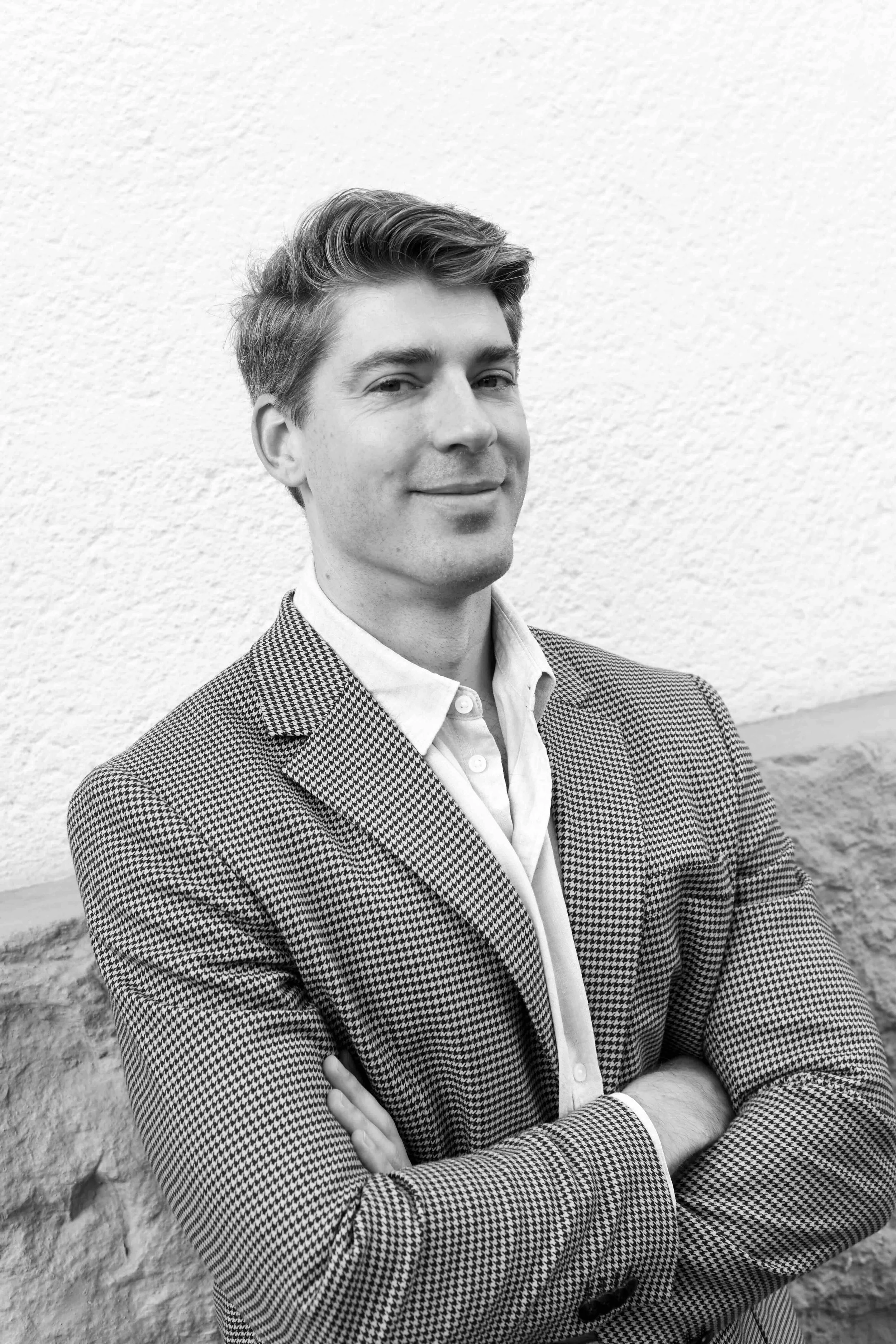 A man with short, styled hair, wearing a checkered blazer over a button-down shirt, standing with arms crossed against a textured wall.
