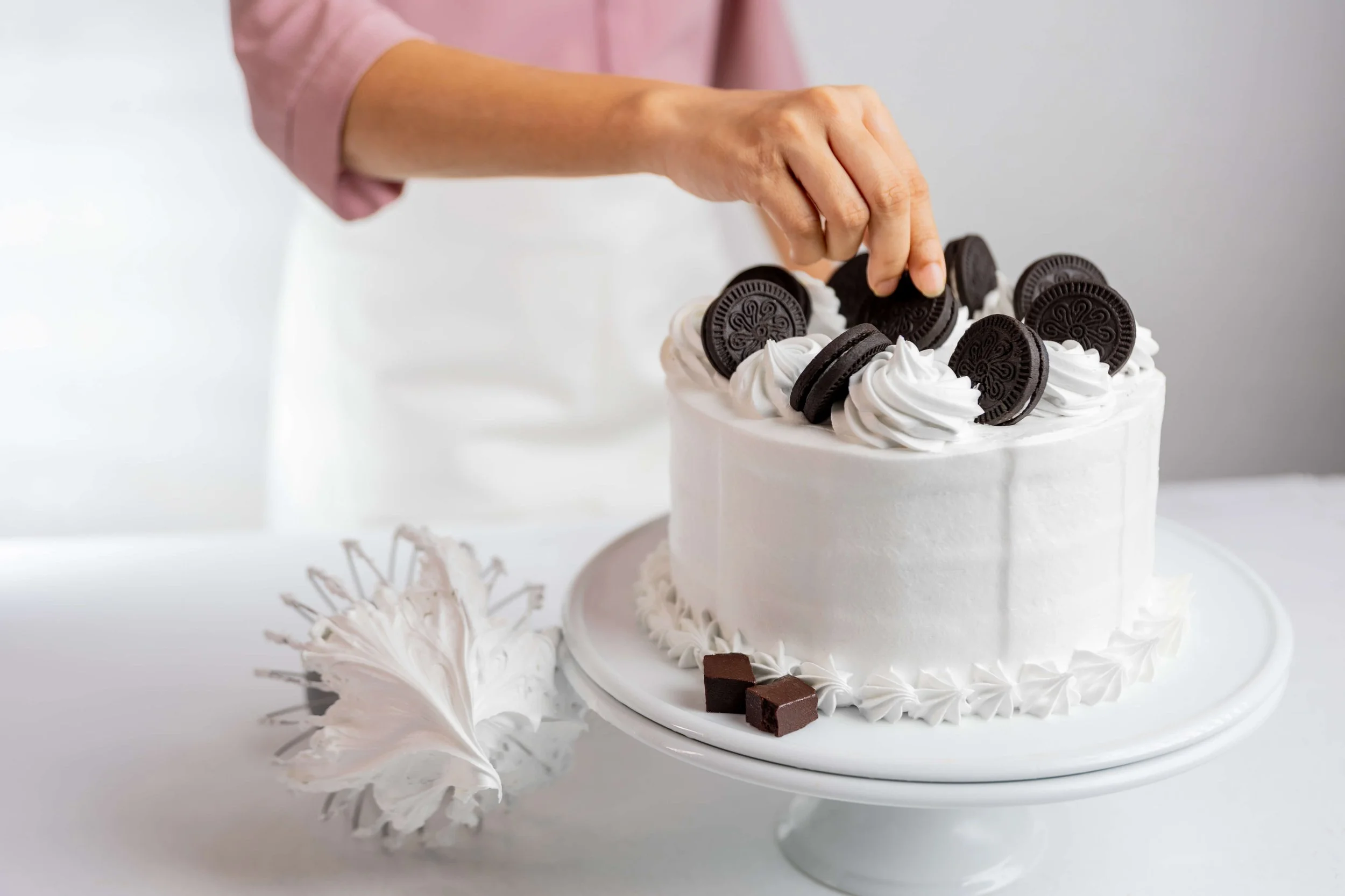 Person decorating a white frosted cake with Oreo cookies and whipped cream, with chocolate truffles nearby