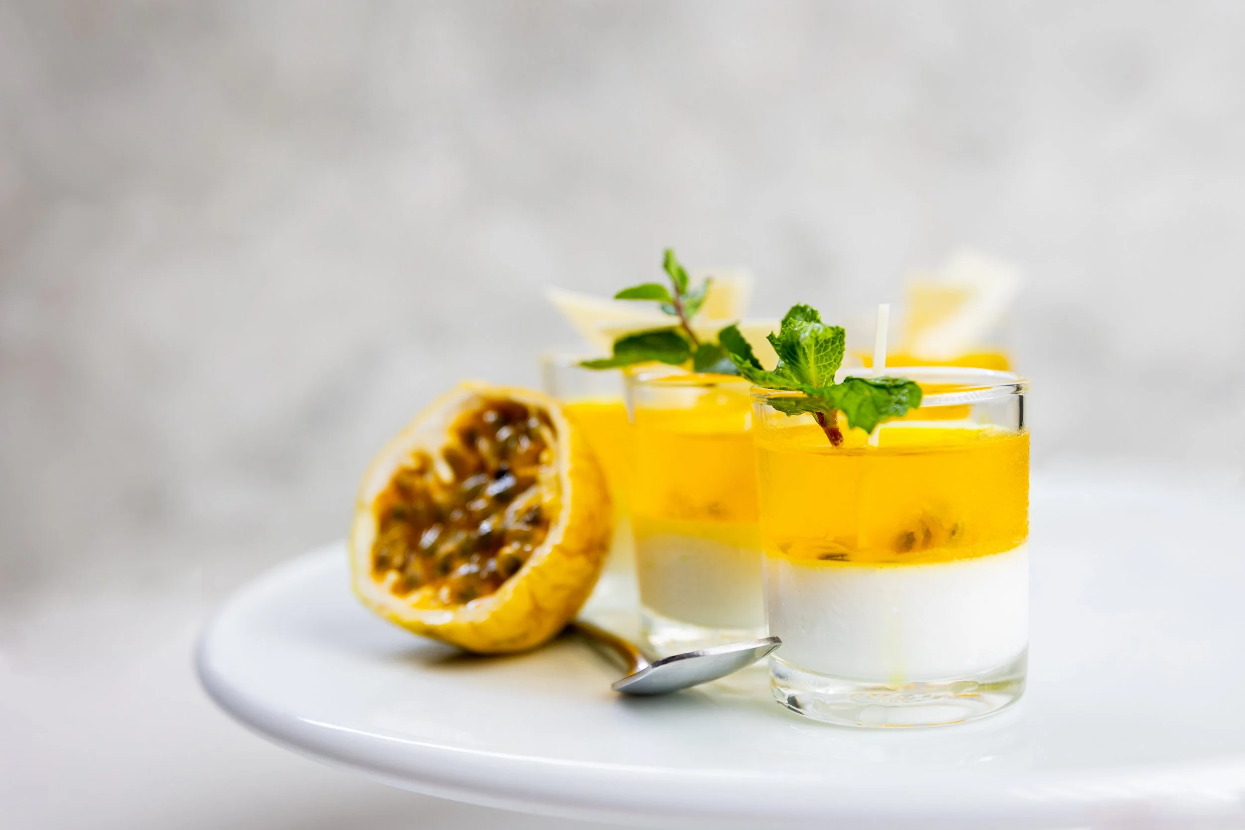 Two layered passion fruit and mango dessert shots garnished with mint leaves and lemon peel, placed on a white plate.