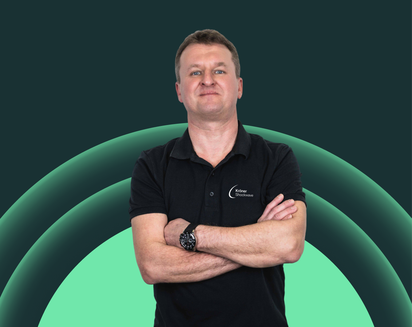 Ein Mann mit gekreuzten Armen trägt ein schwarzes T-Shirt mit dem Logo "Kröner Shockwave" und steht vor einem grafischen Hintergrund mit grünen und dunklen Kreisen.