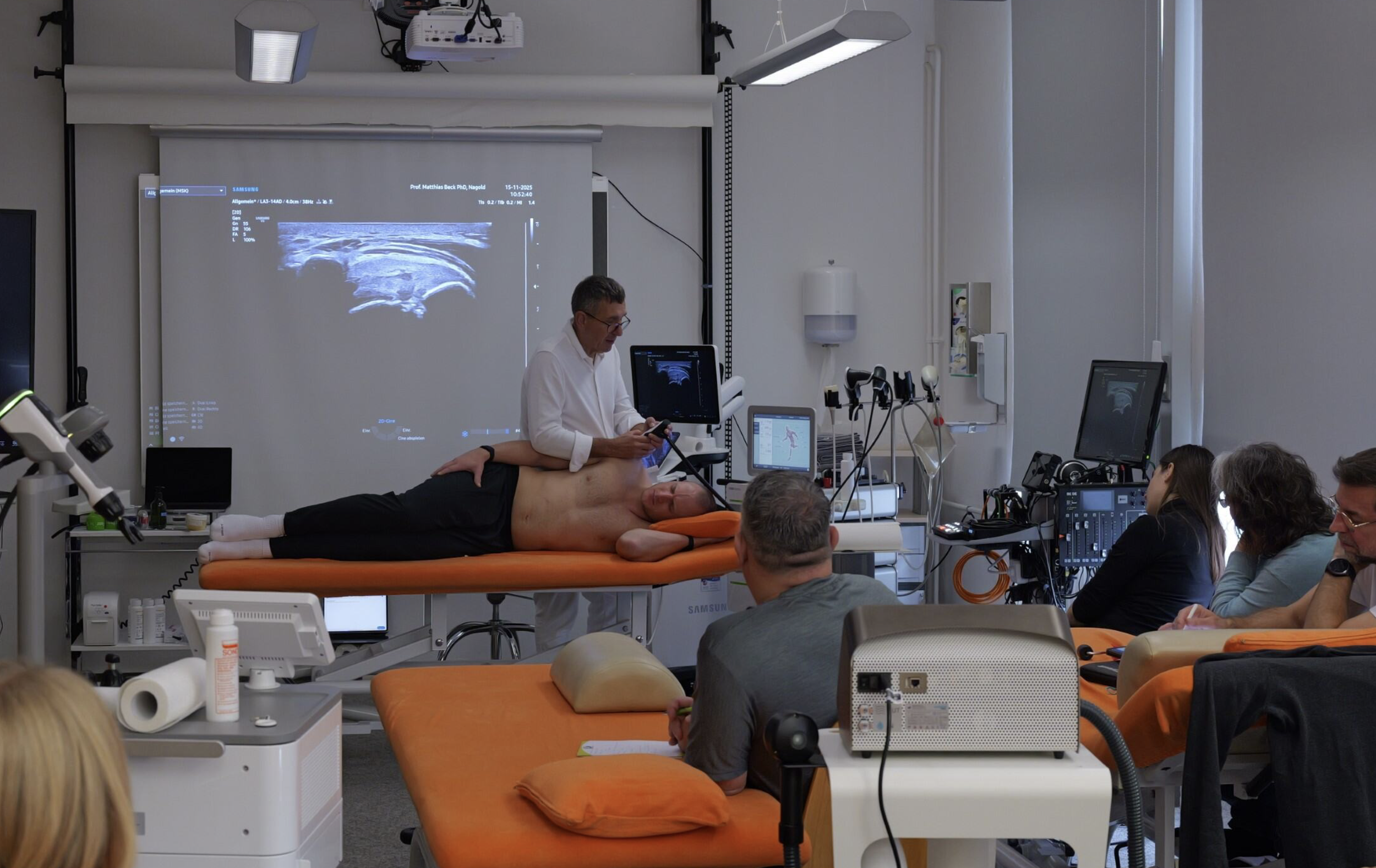 Ein Arzt führt eine Ultraschalluntersuchung an einem liegenden Patienten durch, während andere Personen im Raum zuschauen. Der Raum ist mit medizinischer Ausrüstung und Monitoren ausgestattet.