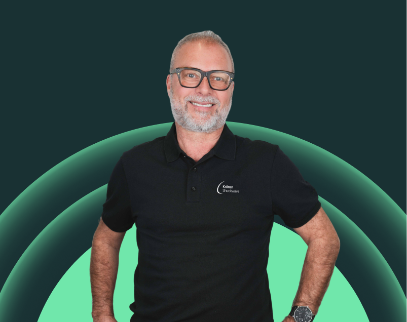Ein Mann mit Brille und grauem Bart, trägt ein schwarzes Polo-Shirt mit einem Logo auf der linken Brustseite, steht vor einem digitalen Hintergrund mit grünen und dunklen Kreisen.
