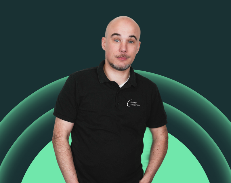 Junger Mann mit kurzen Haaren und Bart, trägt schwarzes Poloshirt, steht vor abstractem grünen Hintergrund.
