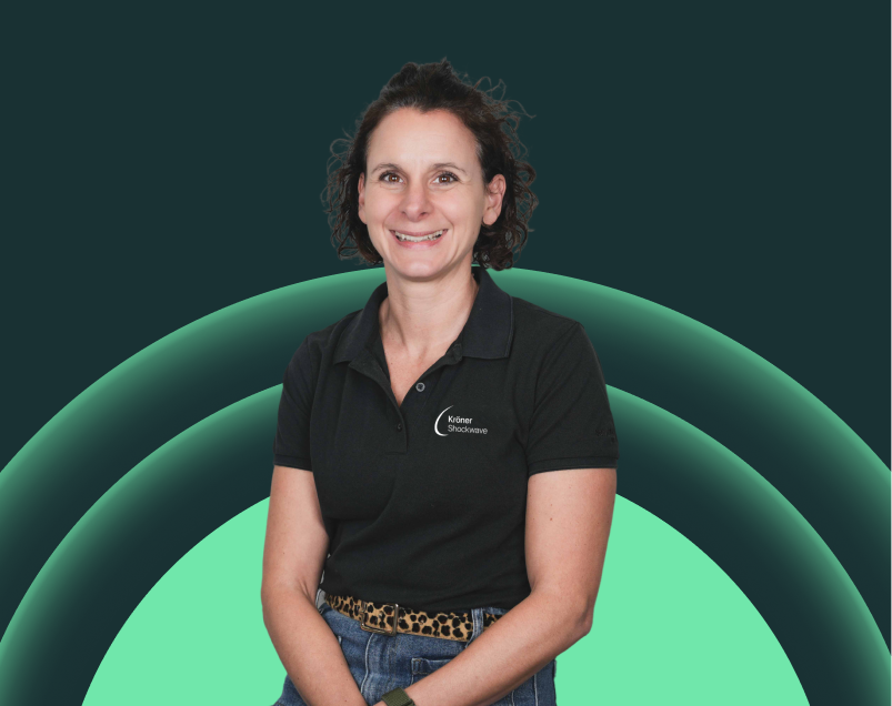 Lächelnde Frau in schwarzen Polo-Shirt mit 'Kräner Shockwave' Logo, vor digitalem Hintergrund mit grünen Bogenlinien.