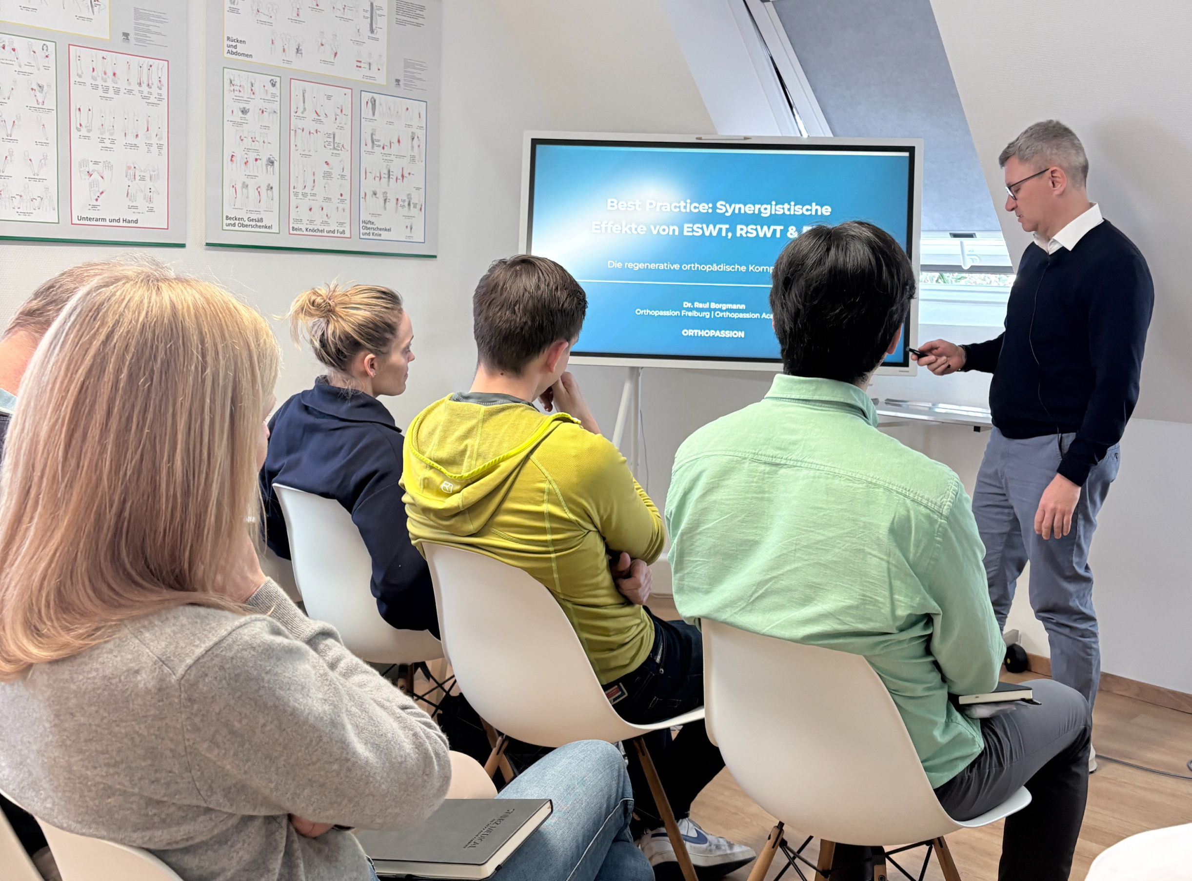 Kroener-Shockwave Mann hält eine Präsentation in einem Klassenraum vor mehreren Zuhörern, während auf einem großen Bildschirm Titel und Name des Referenten zu sehen sind.