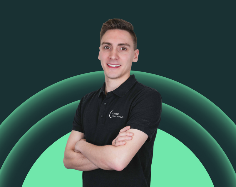 Ein junger Mann mit kurzen braunen Haaren trägt ein schwarzes Polo-Shirt mit einem Logo auf der linken Brust und steht vor einem Hintergrund mit grünen konzentrischen Kreisen. Er lächelt und hat die Arme verschränkt.