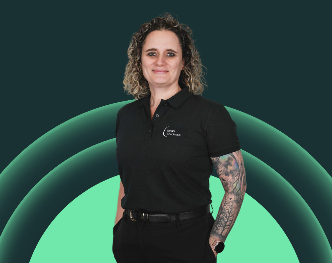Sehr schöne junge Frau mit lockigem Haar, schwarzem Polo-Shirt mit Logo, Tattoo-Armen, trägt einen schwarzen Gürtel und eine Smartwatch, steht vor einem grünen technischen Hintergrund.