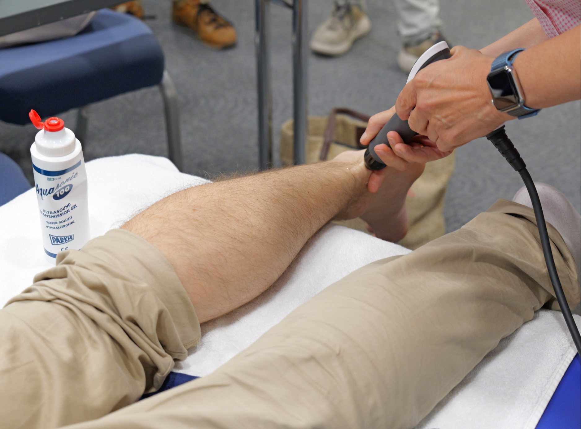 Eine Person liegt auf einer Liege und wird mit einem Ultraschallgerät am Handgelenk untersucht. Es steht ein Fläschchen mit Ultraschall-Gel neben der Liege.