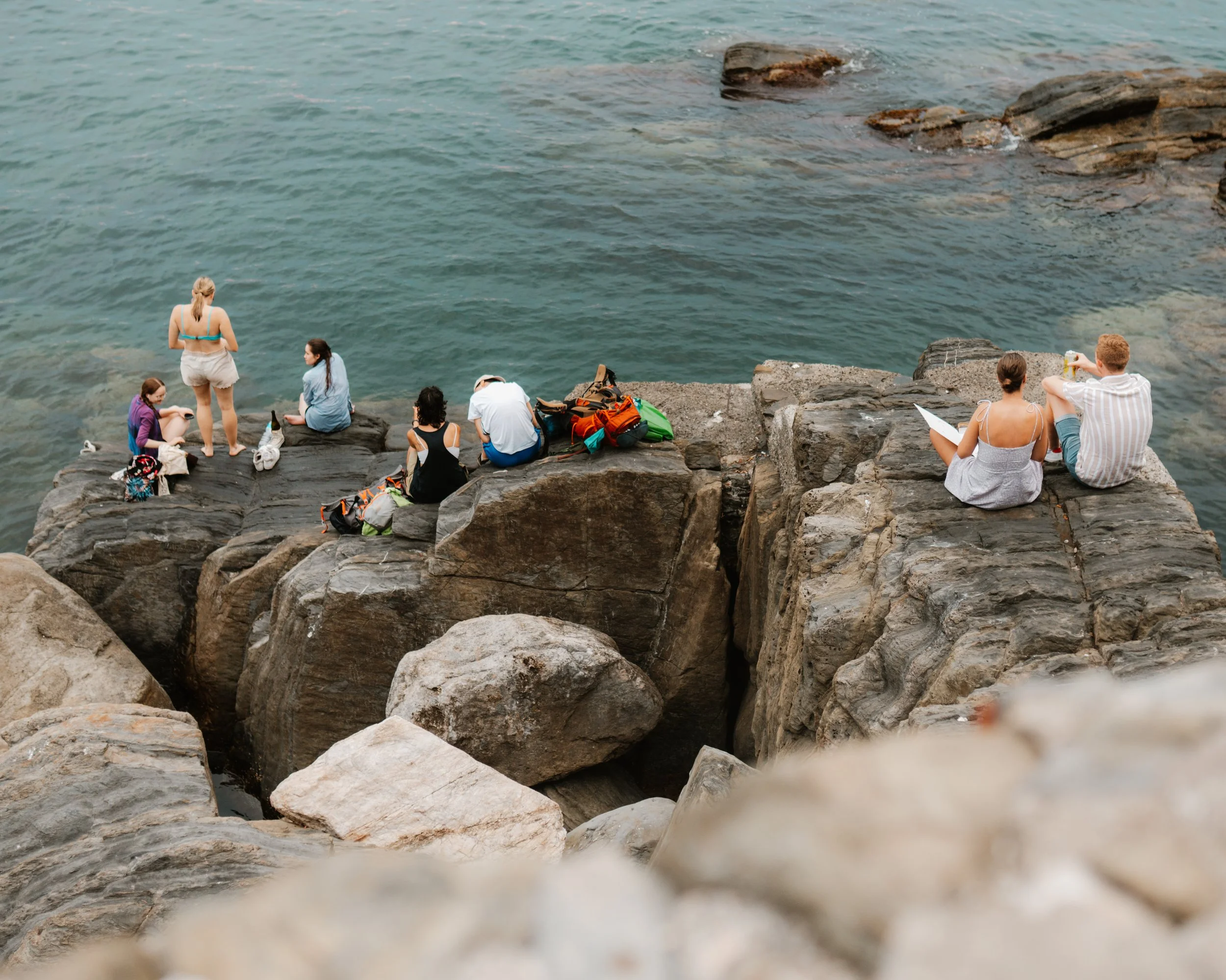 People sitting and relaxing on rocks by the water, some reading, others talking, with bags and drinks nearby.