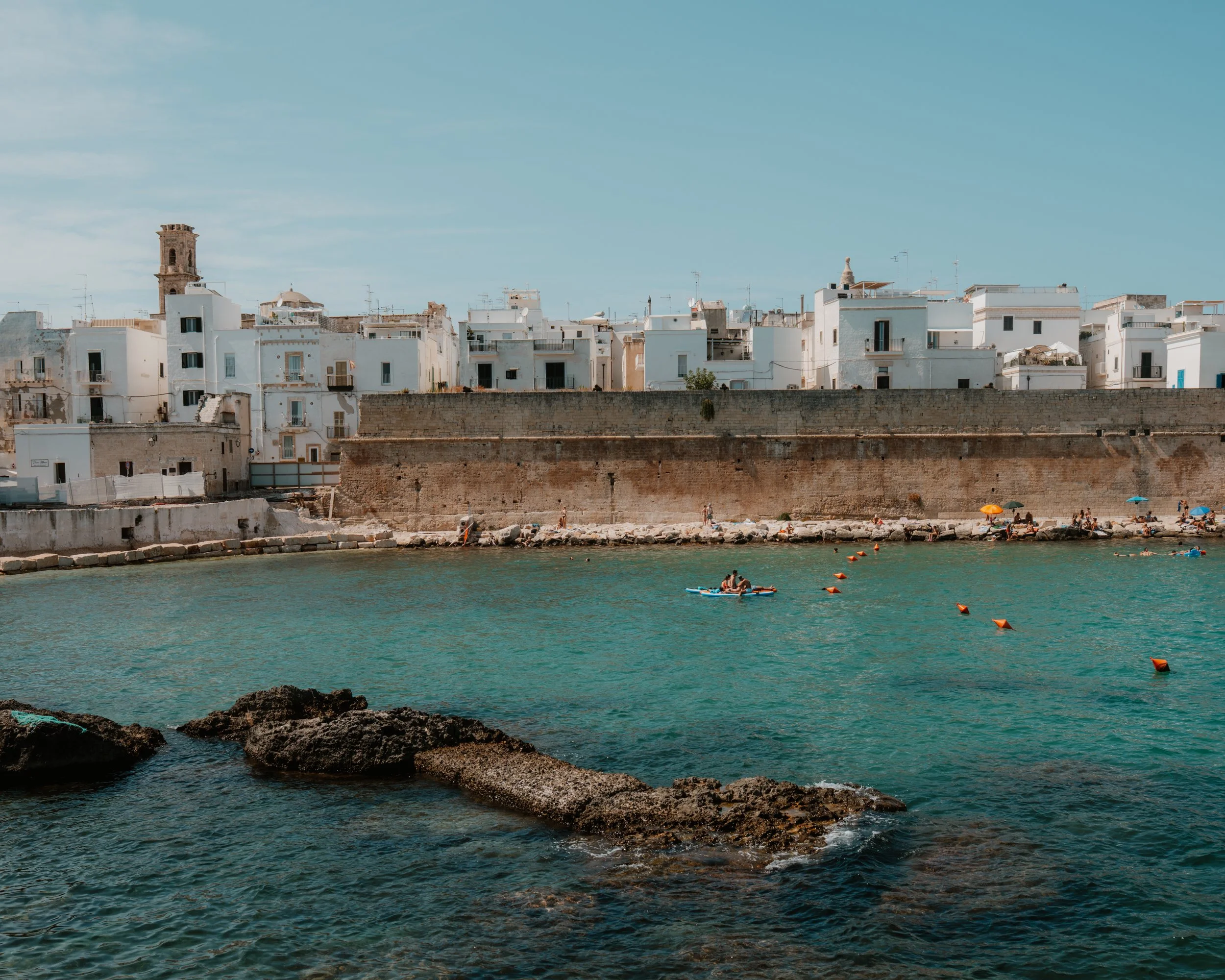 A seaside view of a coastal town with white buildings, a stone wall, and people swimming and relaxing on the beach under colorful umbrellas.