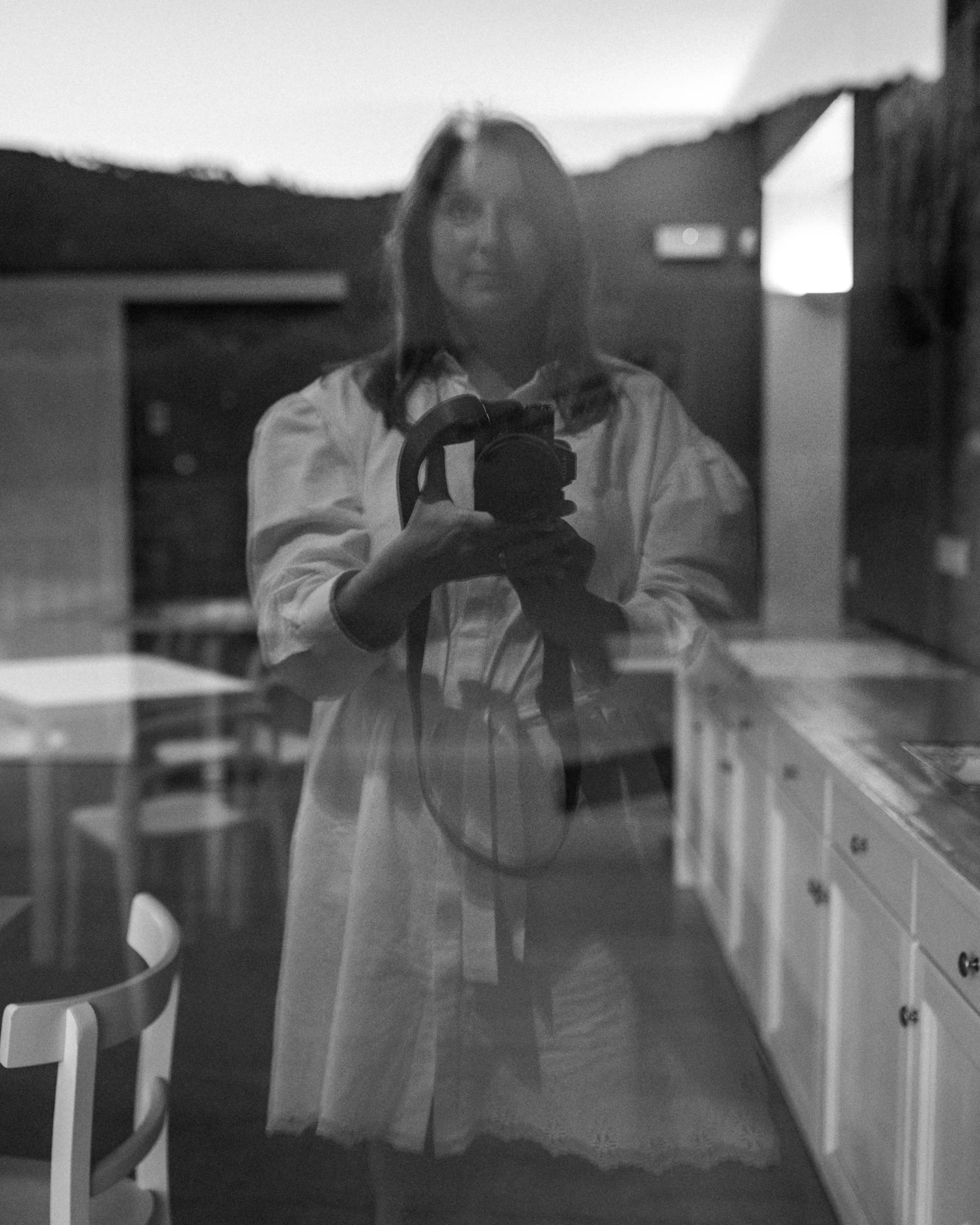Reflection of a woman taking a photo with a camera, standing in a kitchen with cabinets and chairs, in black and white.