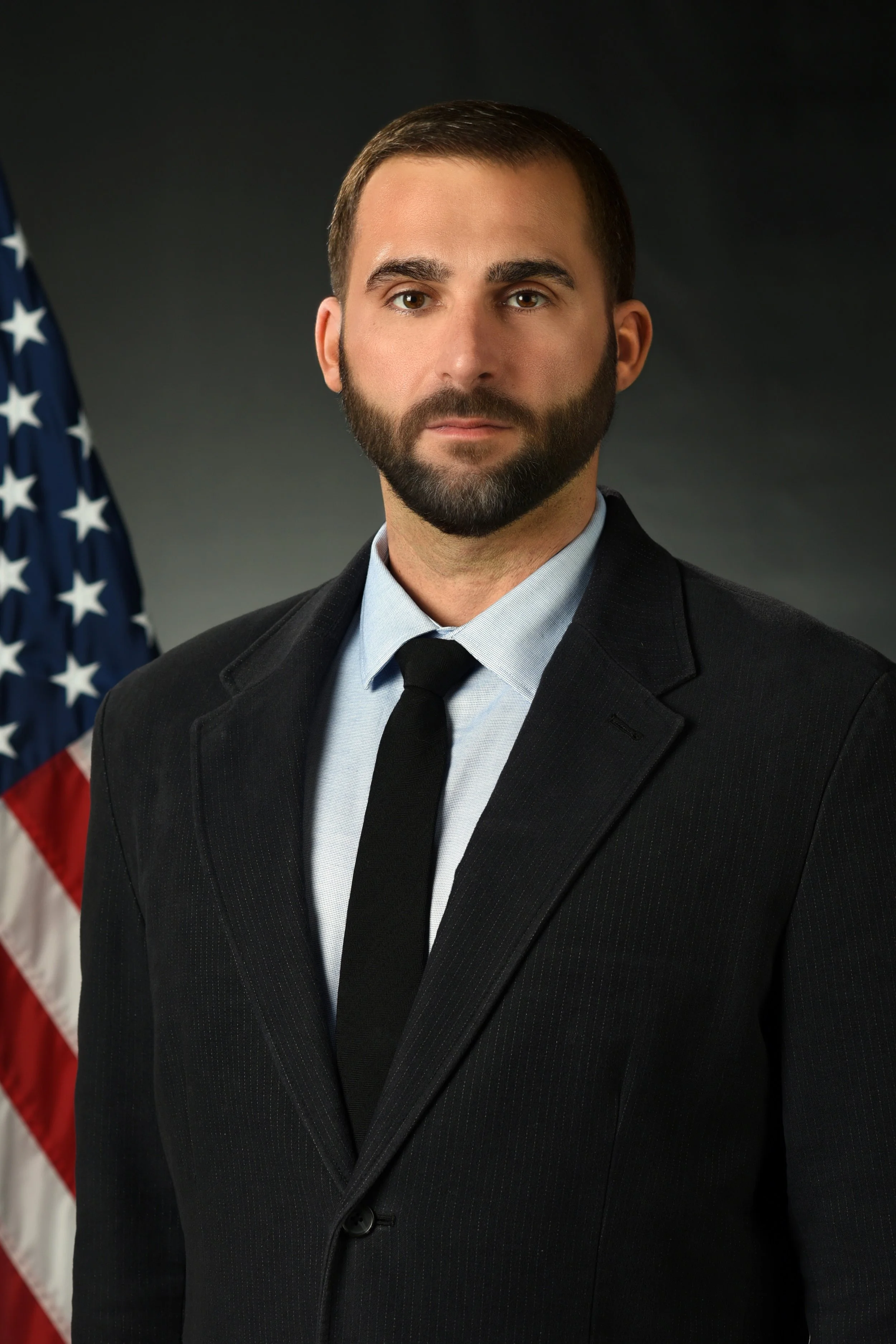 A man in a suit standing in front of the U.S. flag.