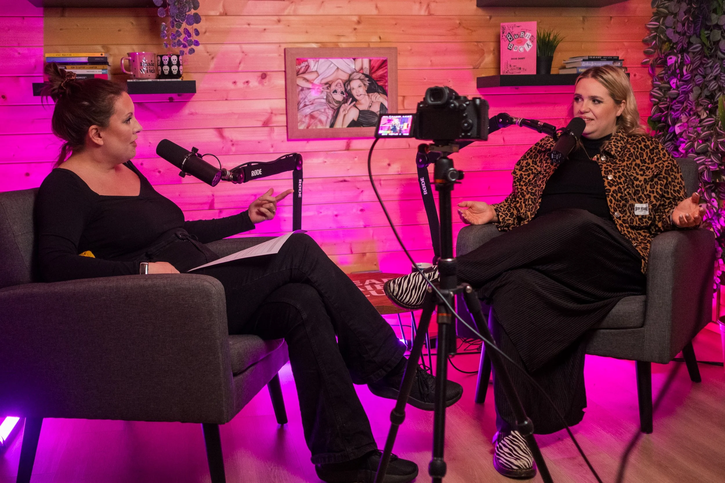 Two women having a conversation on a wedding podcast in a cozy room with pink lighting, wooden wall, and decor including framed artwork, books, and plants.