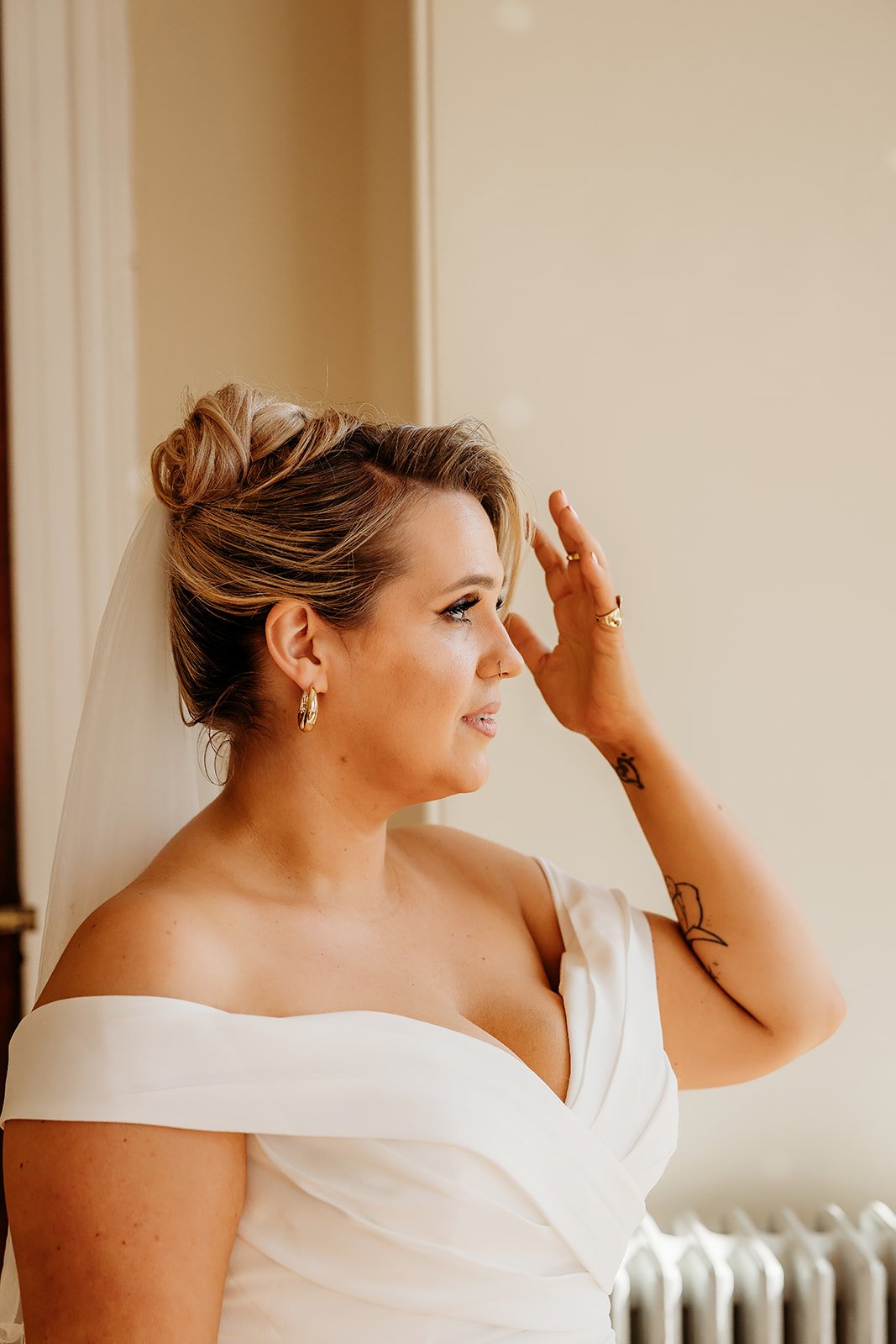 A bride with blonde hair styled in an updo, wearing a white off-the-shoulder wedding dress and gold jewelry, is touching her forehead while looking to the side.