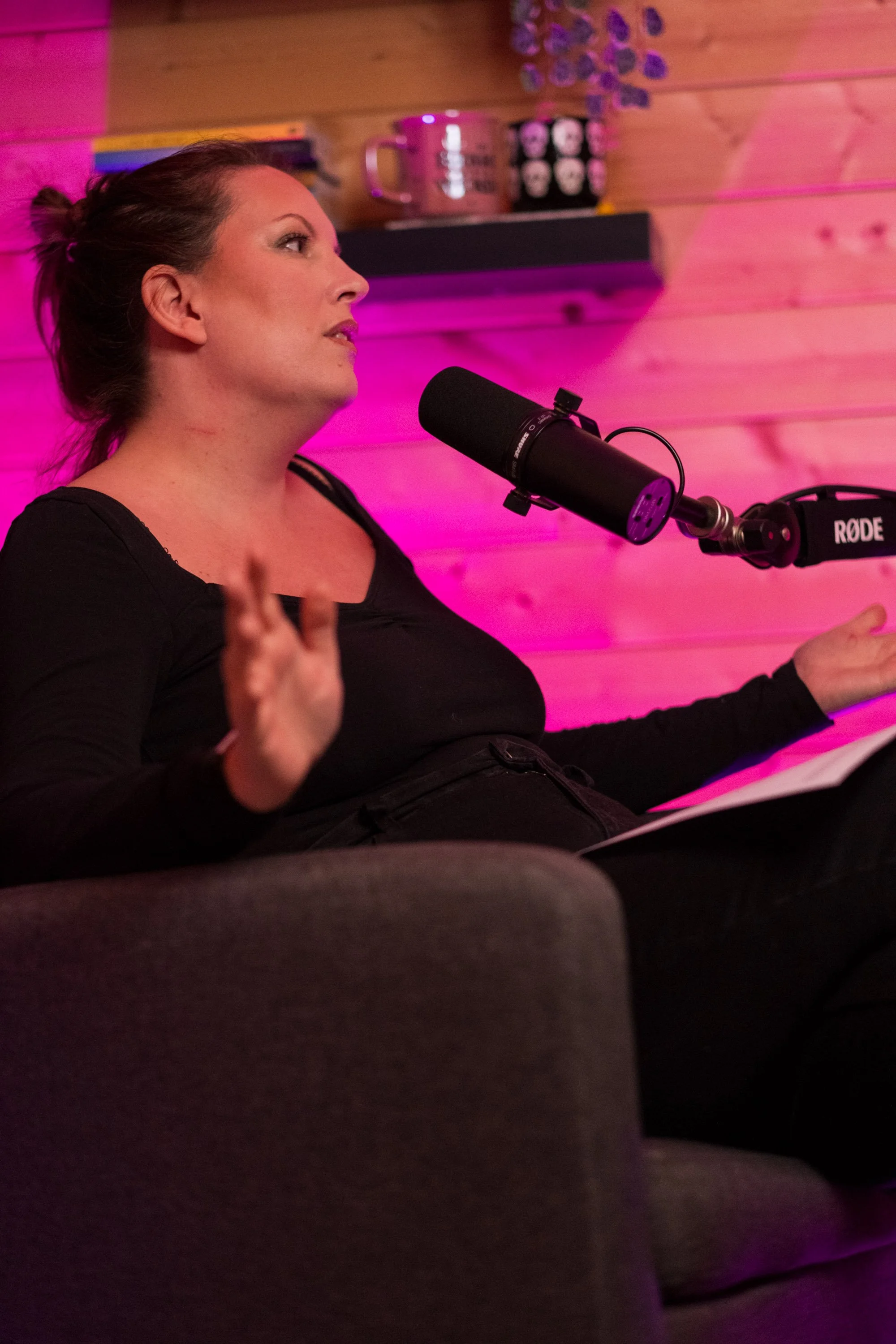 A woman sitting on a couch in a room with pink and purple lighting, speaking into a podcast microphone, with shelves of books, a mug, and decorative items on the wall behind her.
