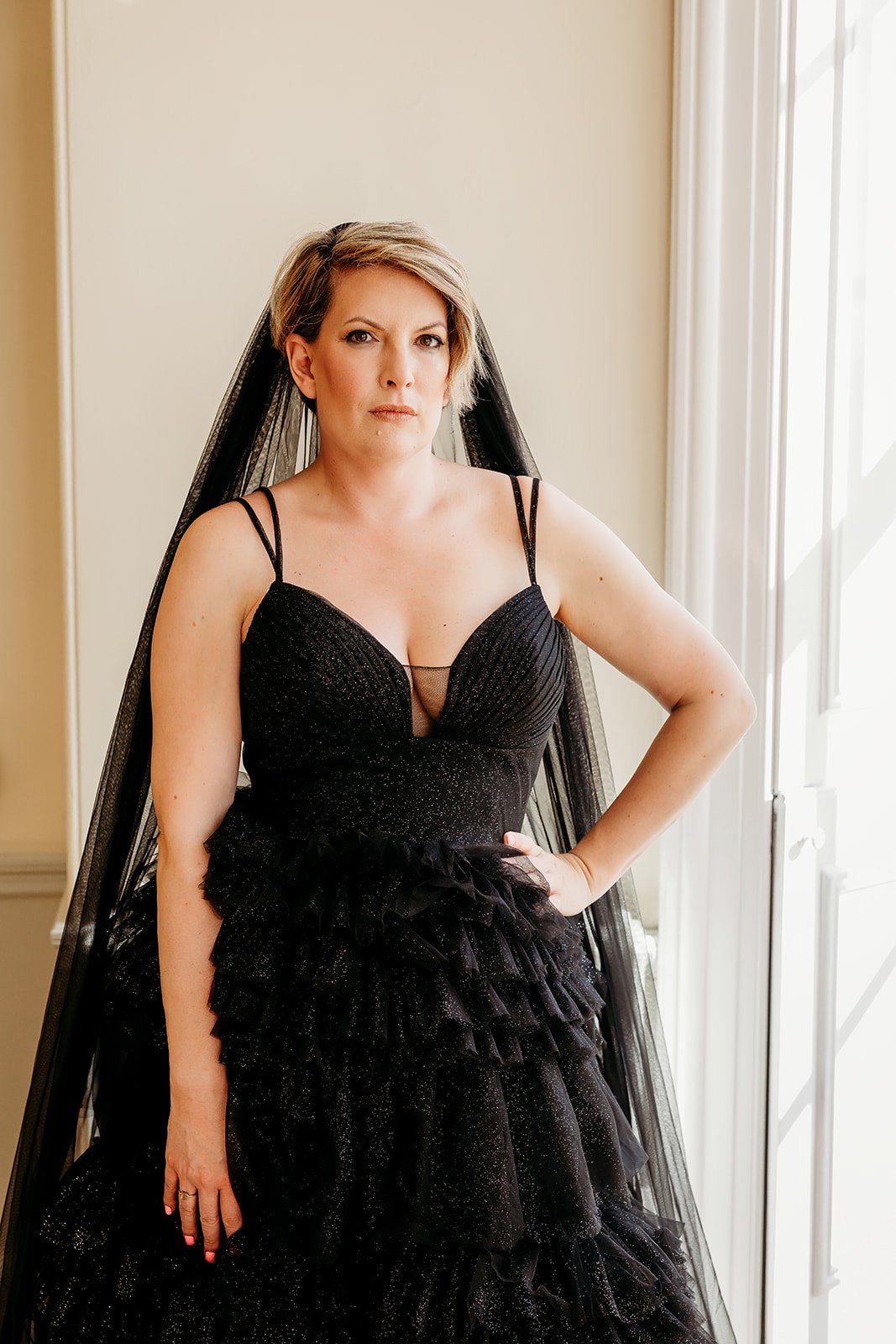 A woman wearing a black wedding dress with spaghetti straps, ruffled skirt, and black veil, standing near a window with light coming in.