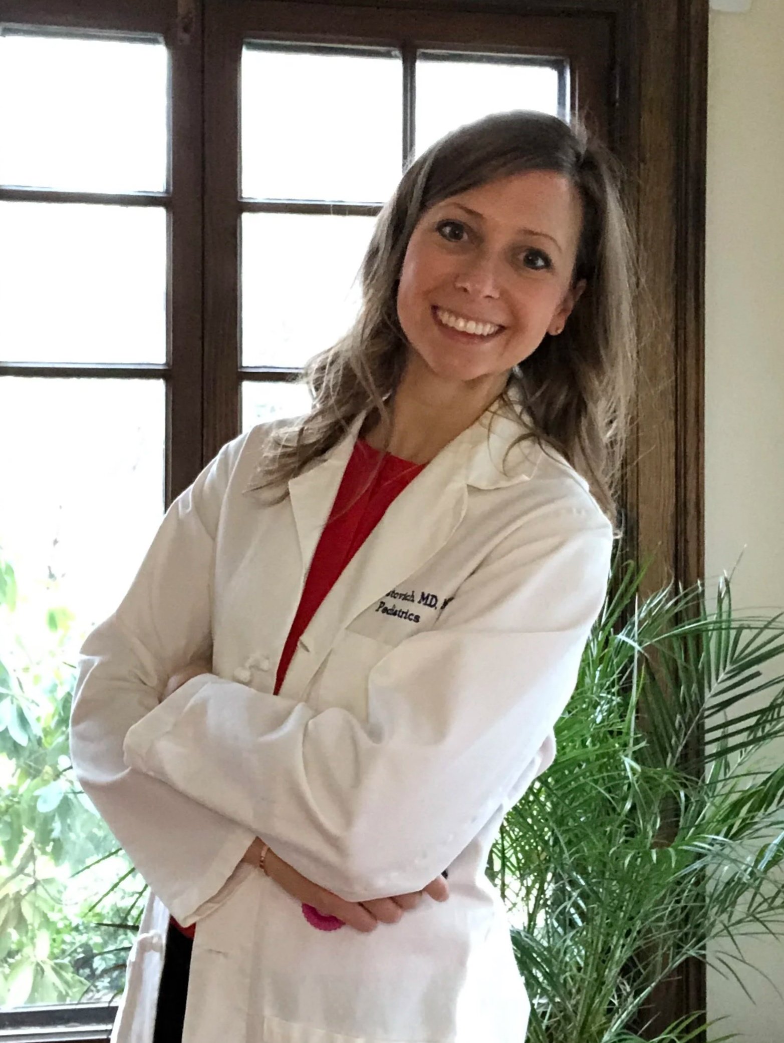 A smiling woman with shoulder-length brown hair wearing a white medical coat standing in front of a window with plants nearby.