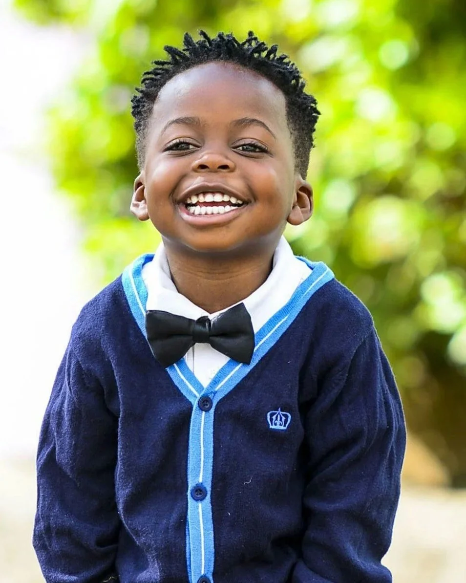 A young boy in a school uniform with a bow tie, smiling outdoors with a background of green foliage.