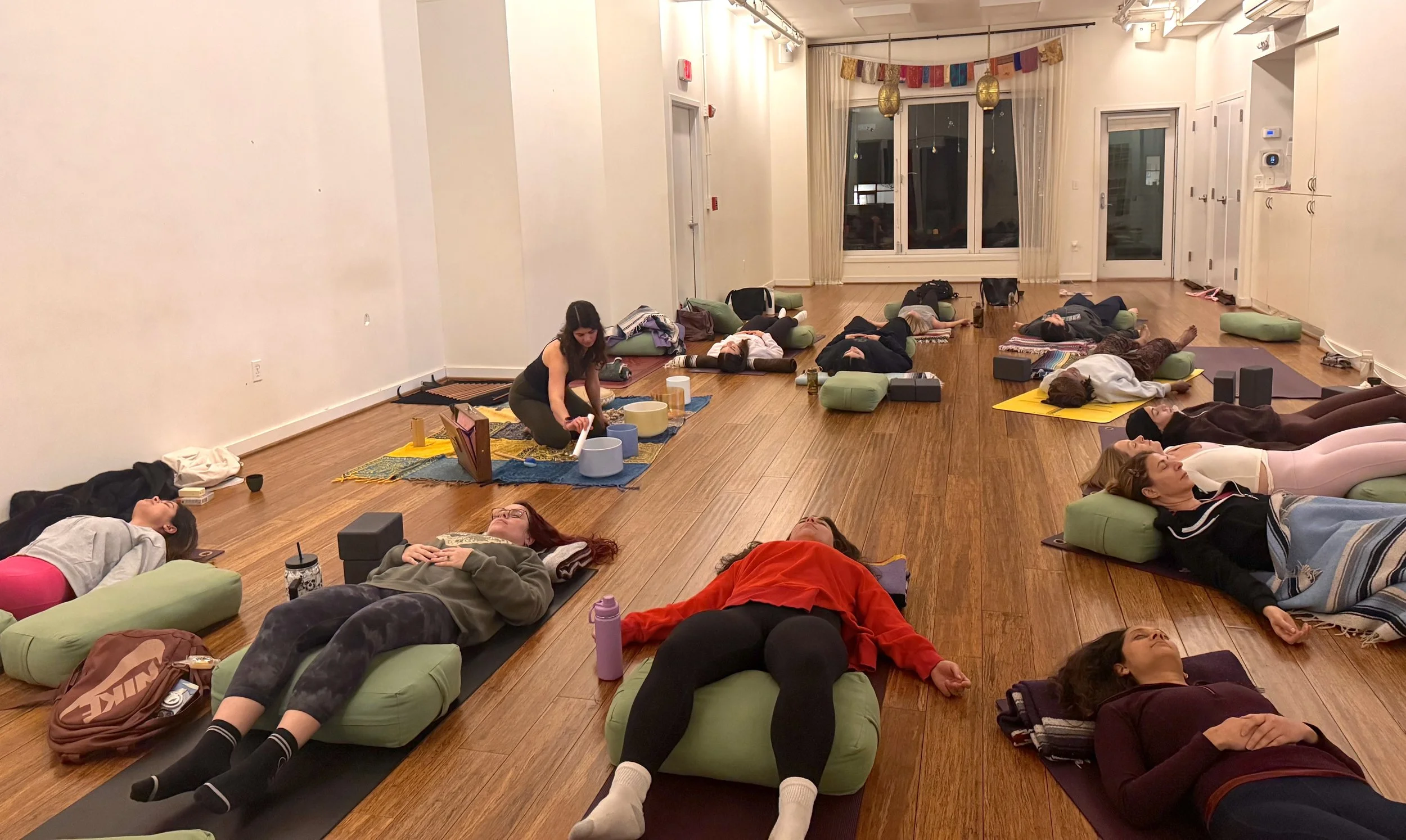 A group of people lying on yoga mats and cushions on a wooden floor, participating in a relaxing sound bath session inside a spacious room with white walls and large windows.