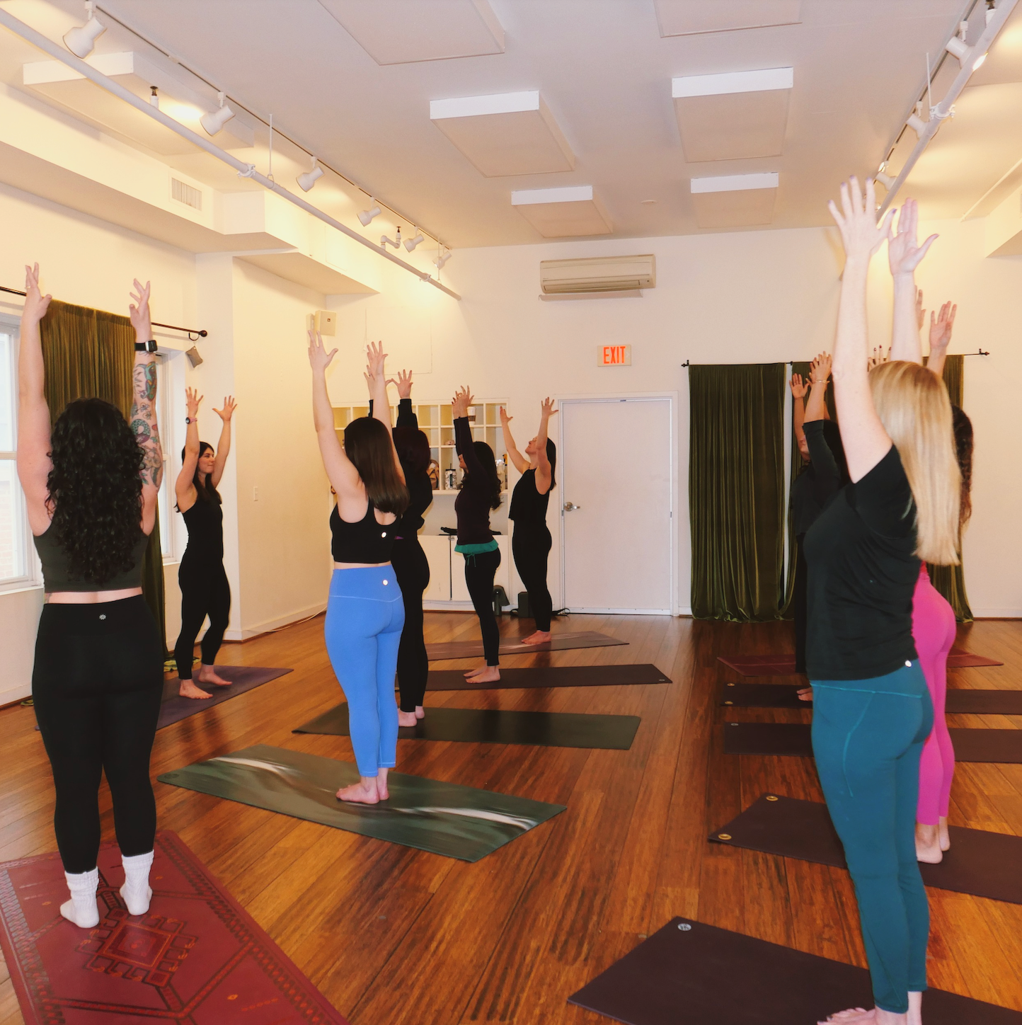 Women practicing yoga in a studio with wood floors and white walls, some with green curtains, and an air conditioner. They are standing on yoga mats, raising their arms upwards in a yoga pose.