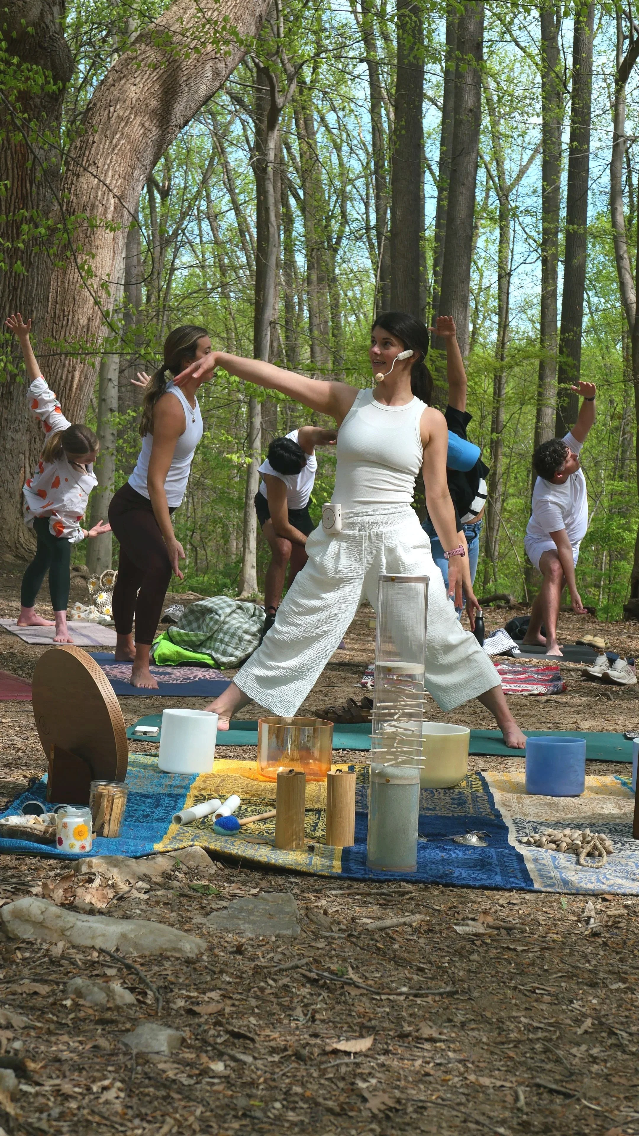 A group of people practicing yoga outdoors in a forest, with a woman leading the session and others following her poses on yoga mats, surrounded by natural elements and various yoga props.