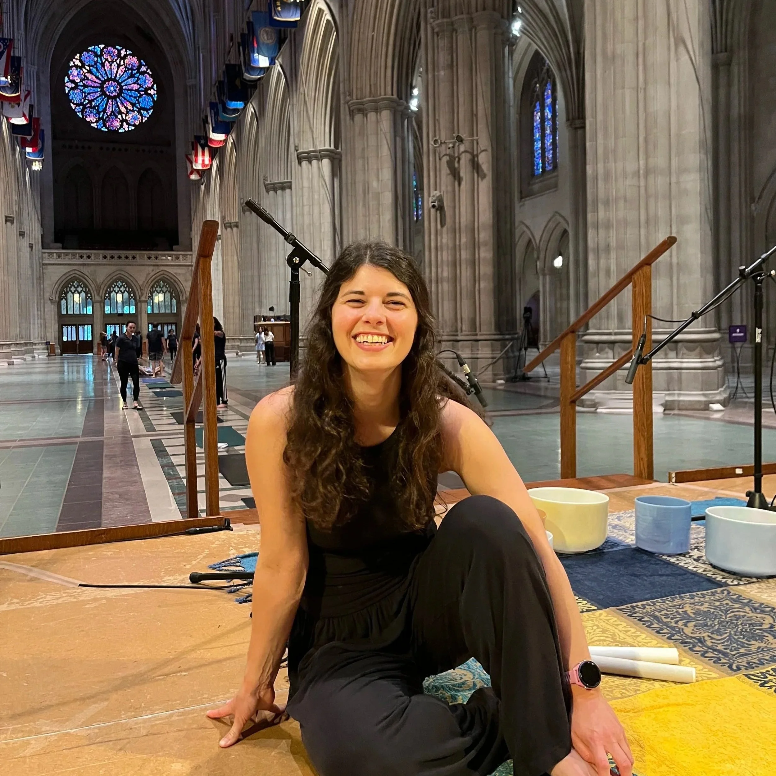 A woman with long, curly hair smiling, practicing yoga in the National Cathedral with high vaulted ceilings, stained glass windows, and flags hanging along the walls.