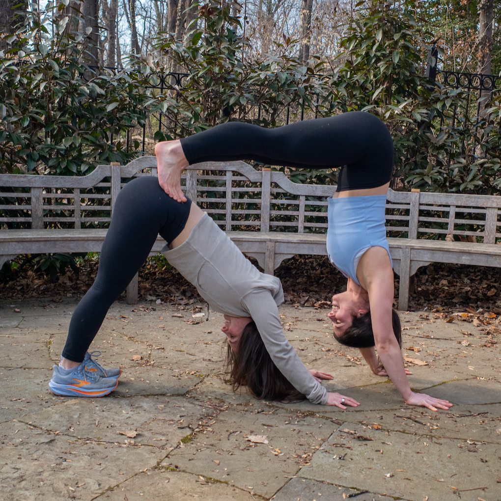 Two women practicing yoga outdoors in a backyard, forming a double bridge pose with their bodies. One woman is wearing a gray hoodie and black leggings, while the other is in a blue tank top and black leggings. They are smiling and facing each other, with the woman in the gray hoodie supporting the other upside down.