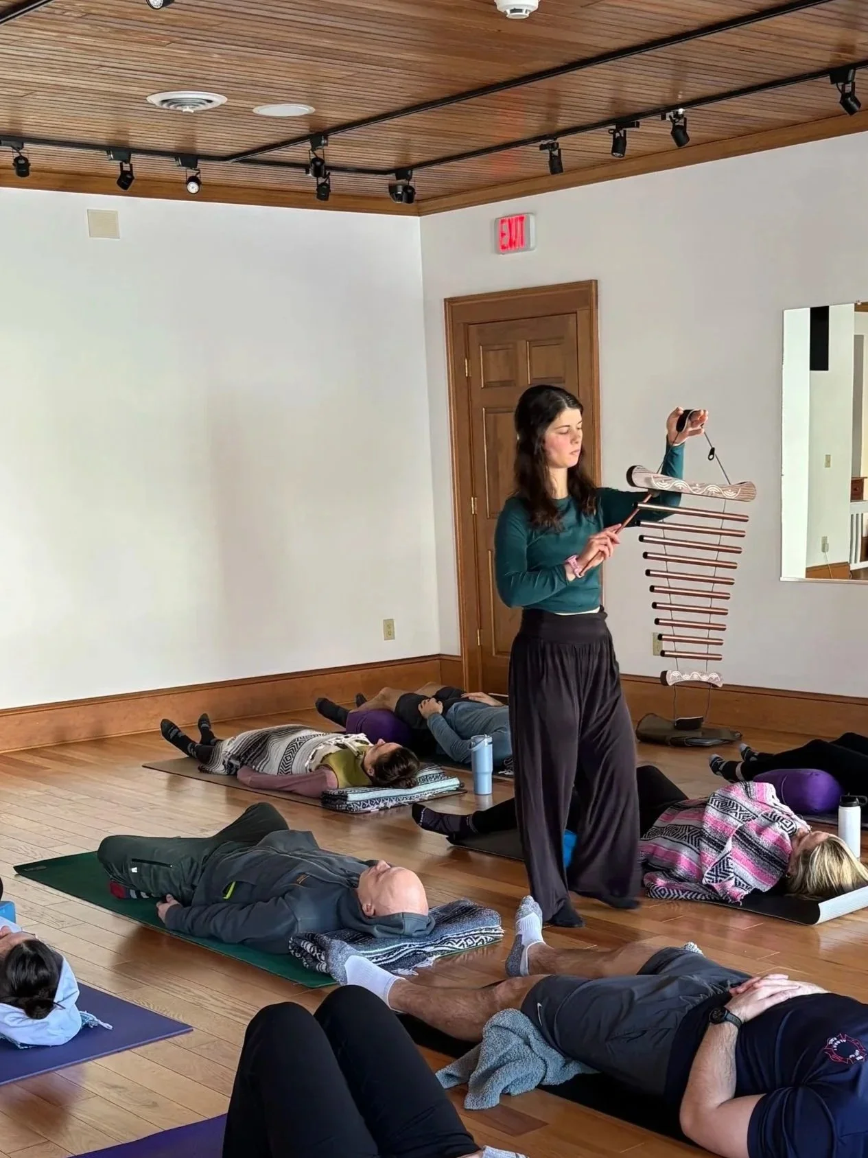 A yoga class taking place in a room with wooden floors and ceiling. Participants are lying on mats in a relaxation pose, while an instructor in the center holds meditation chimes, leading the session.