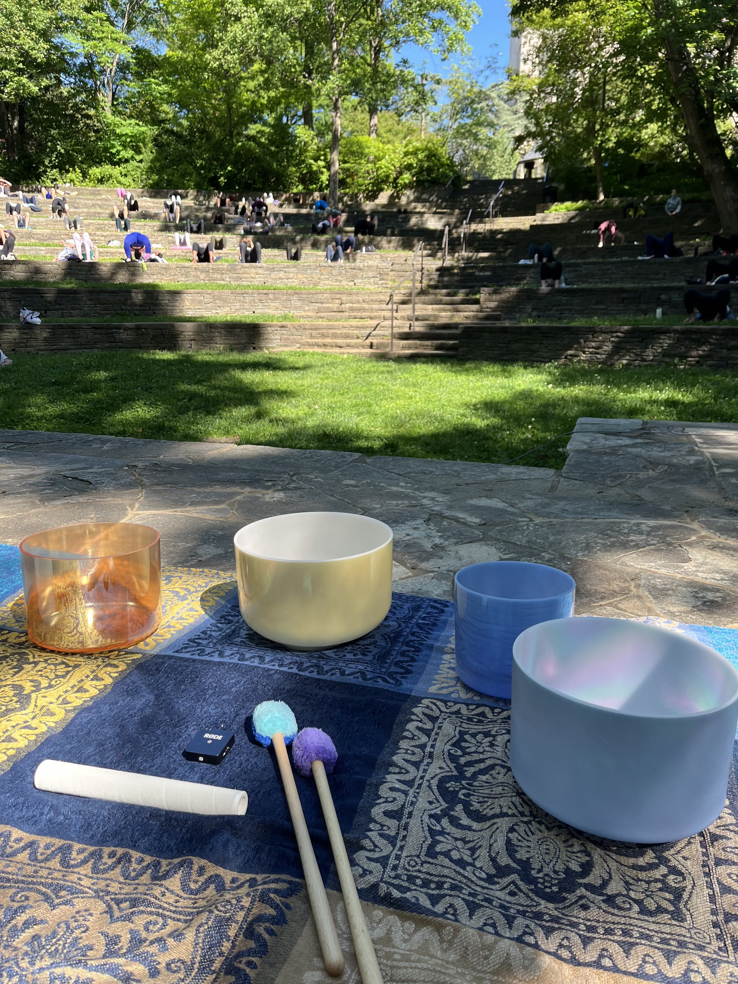 A view from a stone table showing crystal alchemy singing bowls, a purple pom-pom mallet, and a white stick, with an outdoor amphitheater filled with people doing stretches or yoga in the background, under trees on a sunny day.