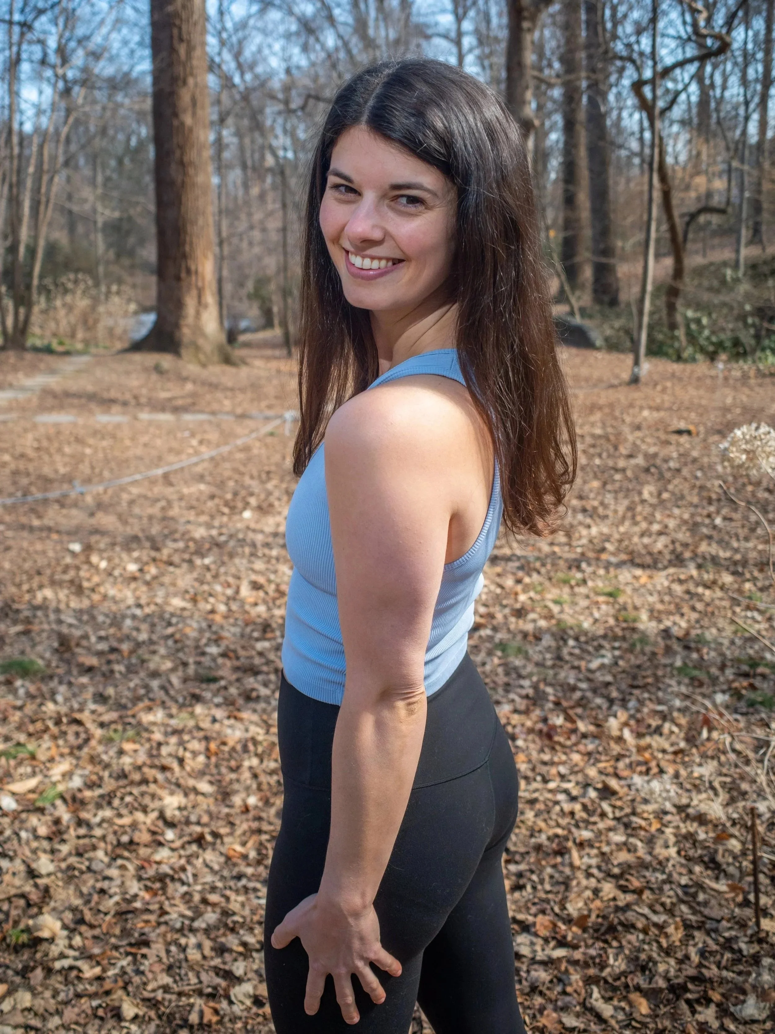 A woman with long, dark hair smiling and looking at the camera, standing outdoors in a wooded area with many trees and fallen leaves on the ground.