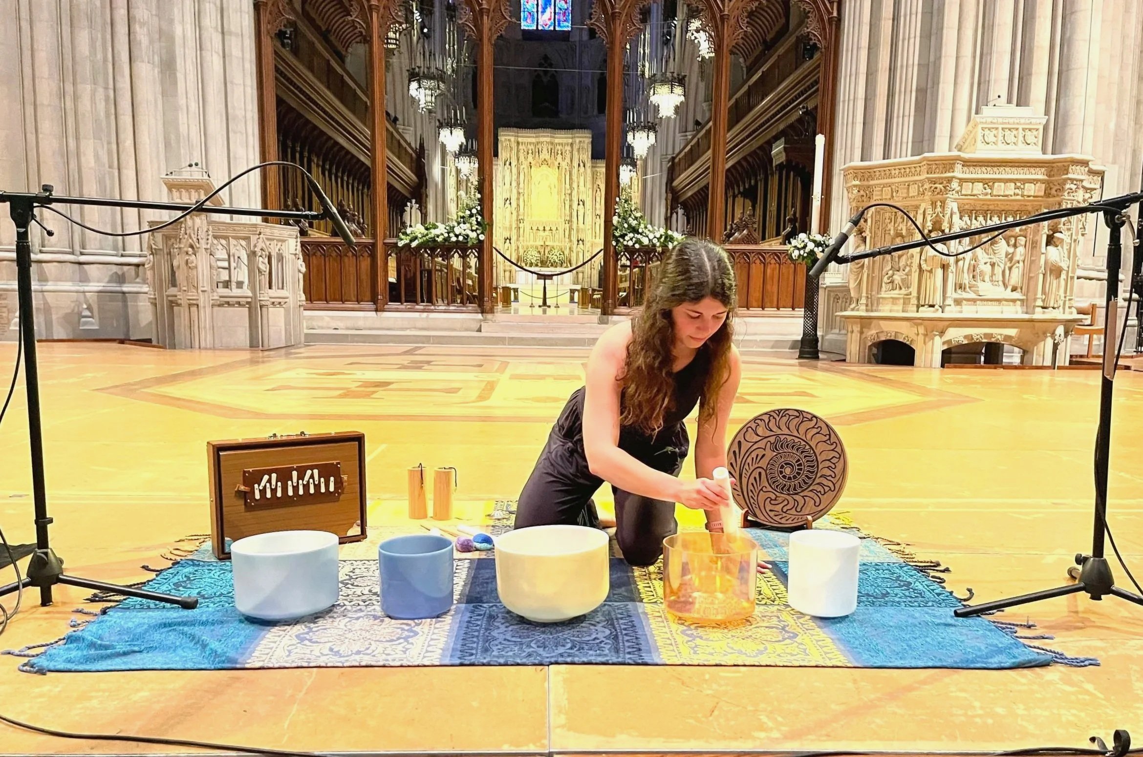 A young woman kneeling on a blue patterned rug inside the National Cathedral, surrounded by musical instruments and crystal singing bowls, with microphones on stands, and a backdrop of ornate church architecture and stained glass windows.