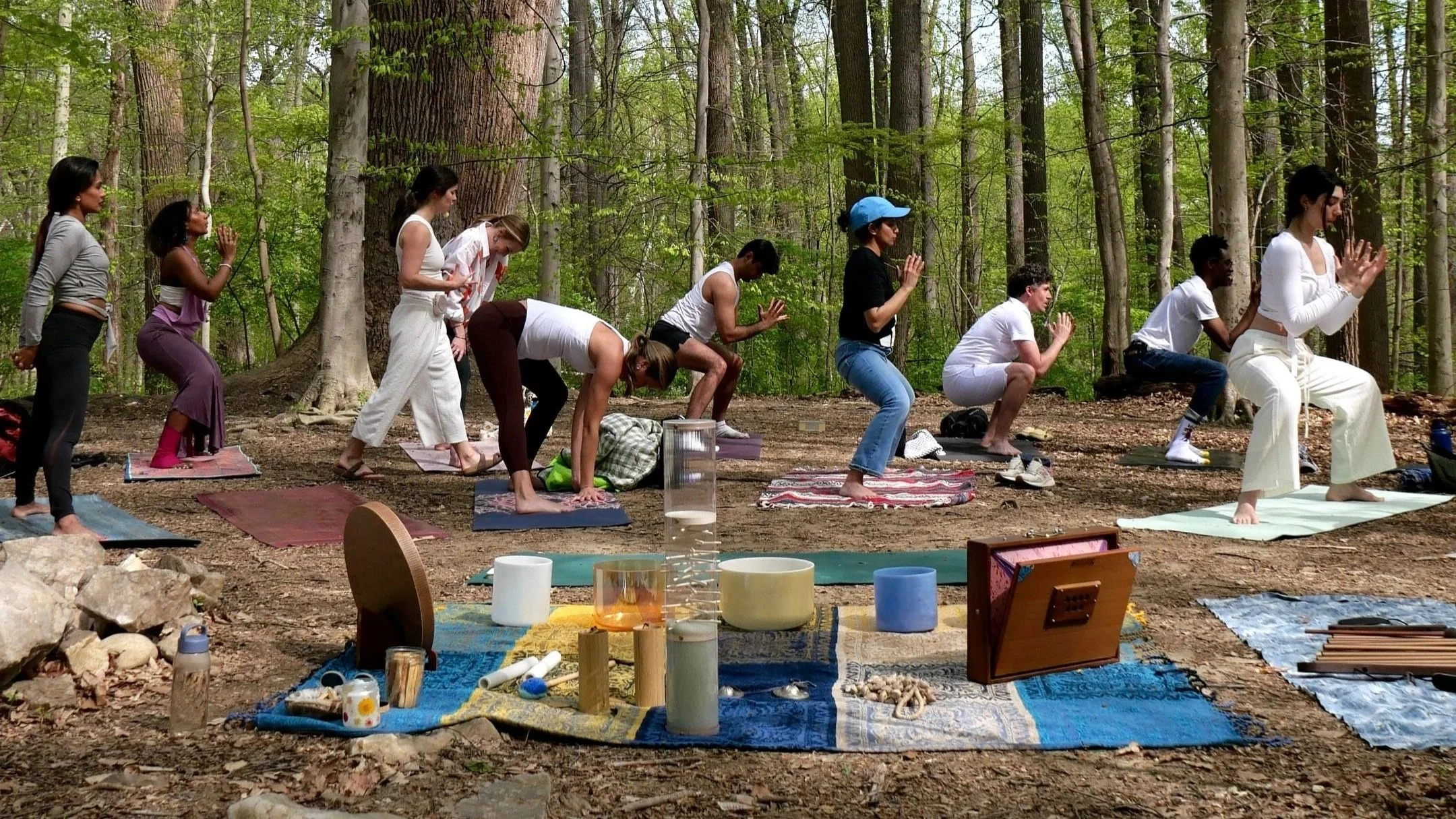 Group of people practicing yoga outdoors in a forest, standing on yoga mats with their hands in prayer position, surrounded by trees and forest floor with various objects and cushions.