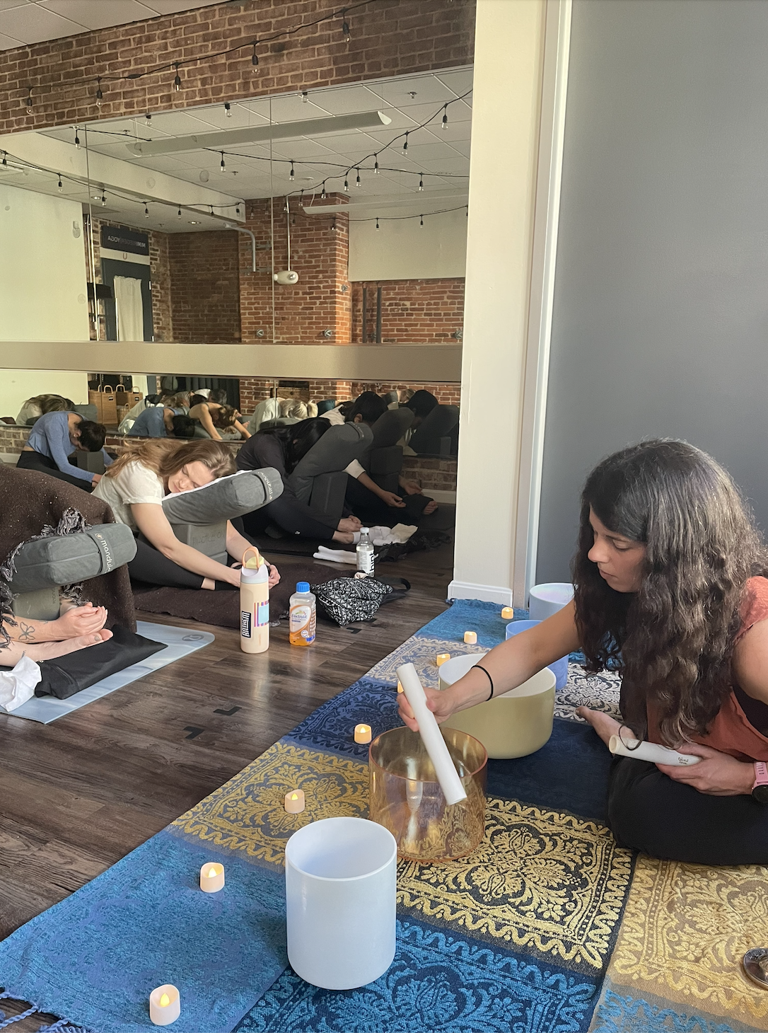Group of people practicing yoga and experiencing a sound bath in a studio with exposed brick walls and string lights, seated on mats and chairs, with a facilitator using singing bowls.