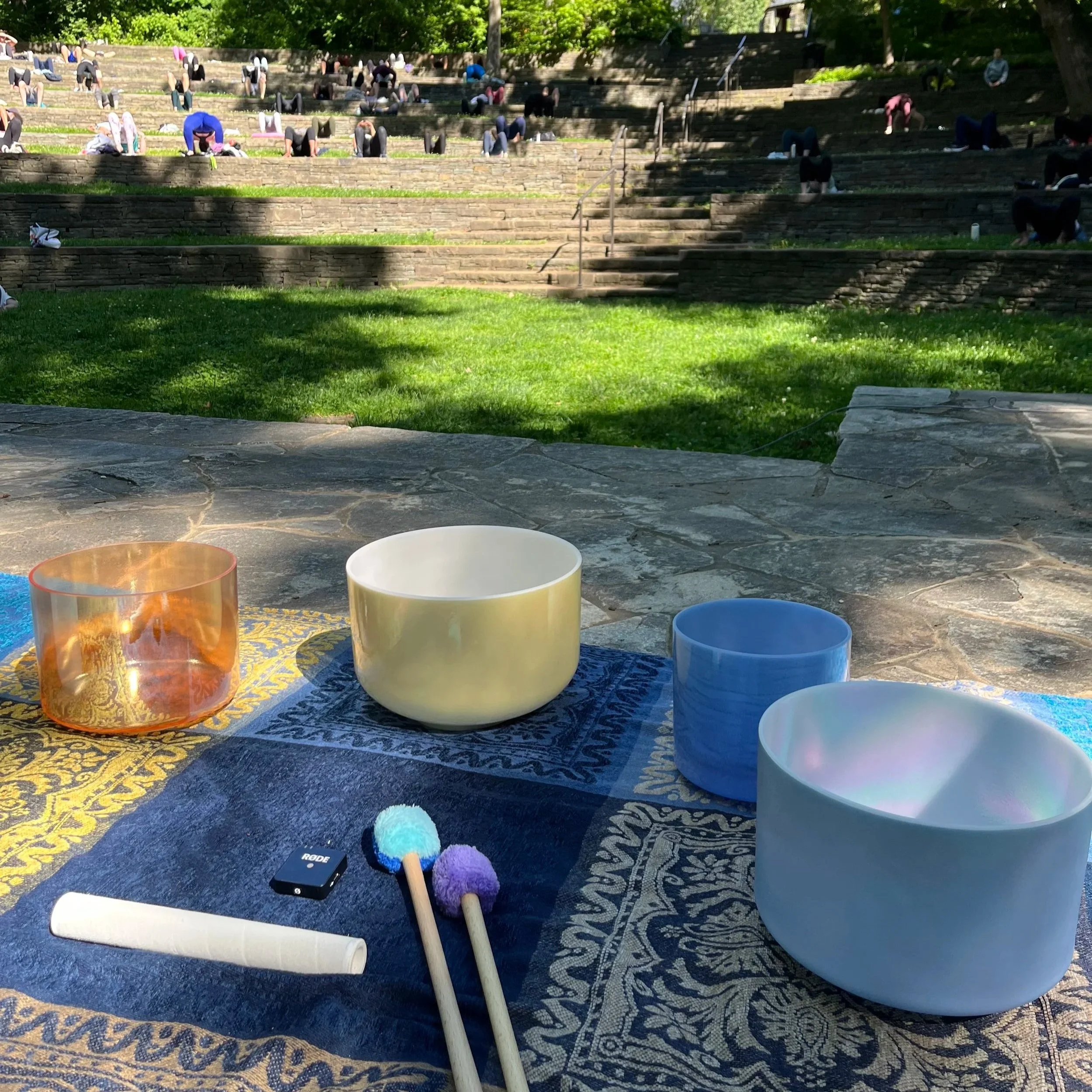 Outdoor scene of tiered stone steps with people practicing yoga, and a foreground with various colorful crystal alchemy singing bowls and small percussion instruments on a patterned rug.