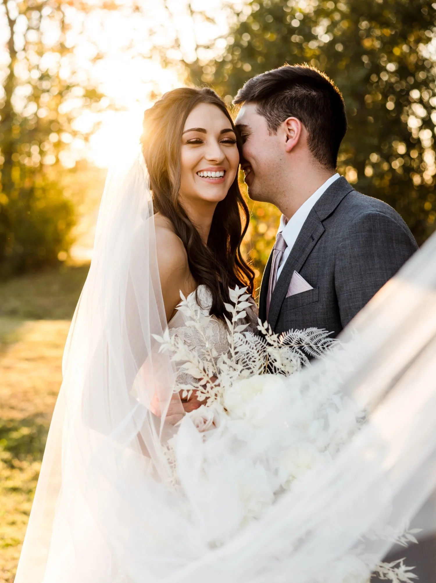 A happy bride and groom smiling and sharing a private moment outdoors during sunset, with trees in the background.